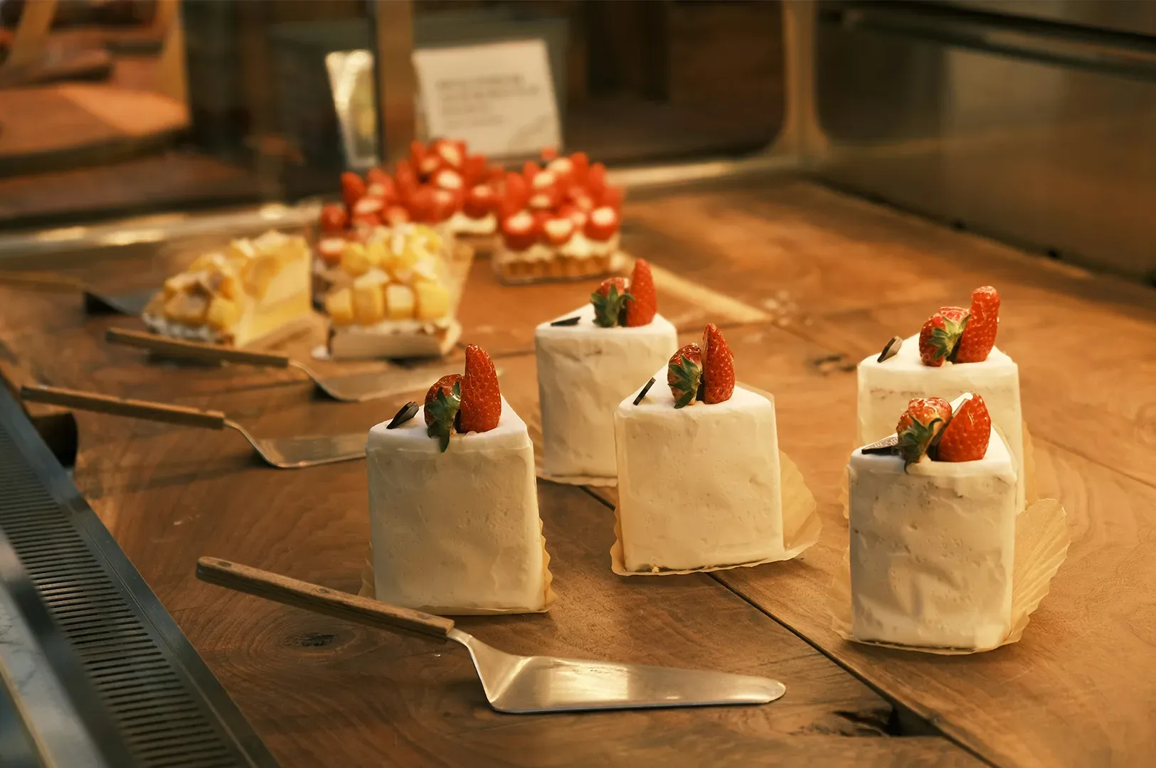 Mini white dessert cakes topped with strawberries on a wooden buffet table, with tongs nearby