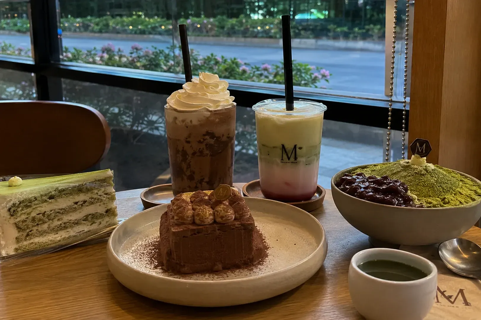 Dessert table with cake slices, layered drinks, and a bowl of green tea dessert by a window.