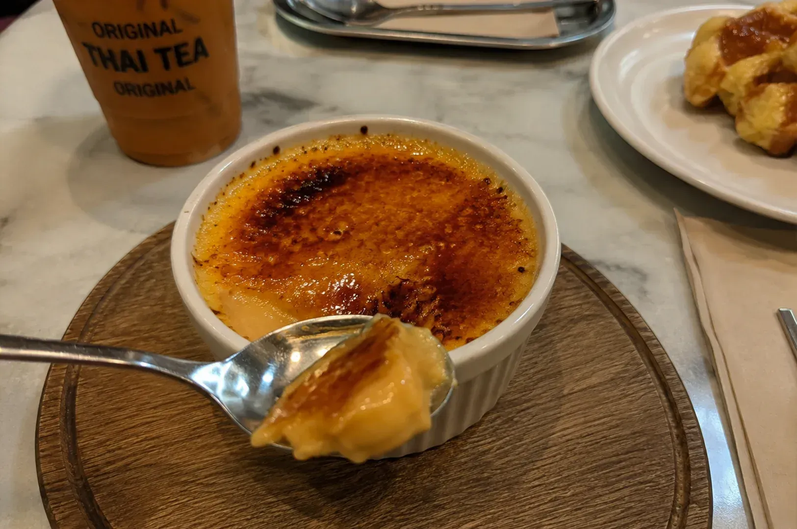 Crème brûlée with a spoon on a woven placemat, with tea and pastries in the background.