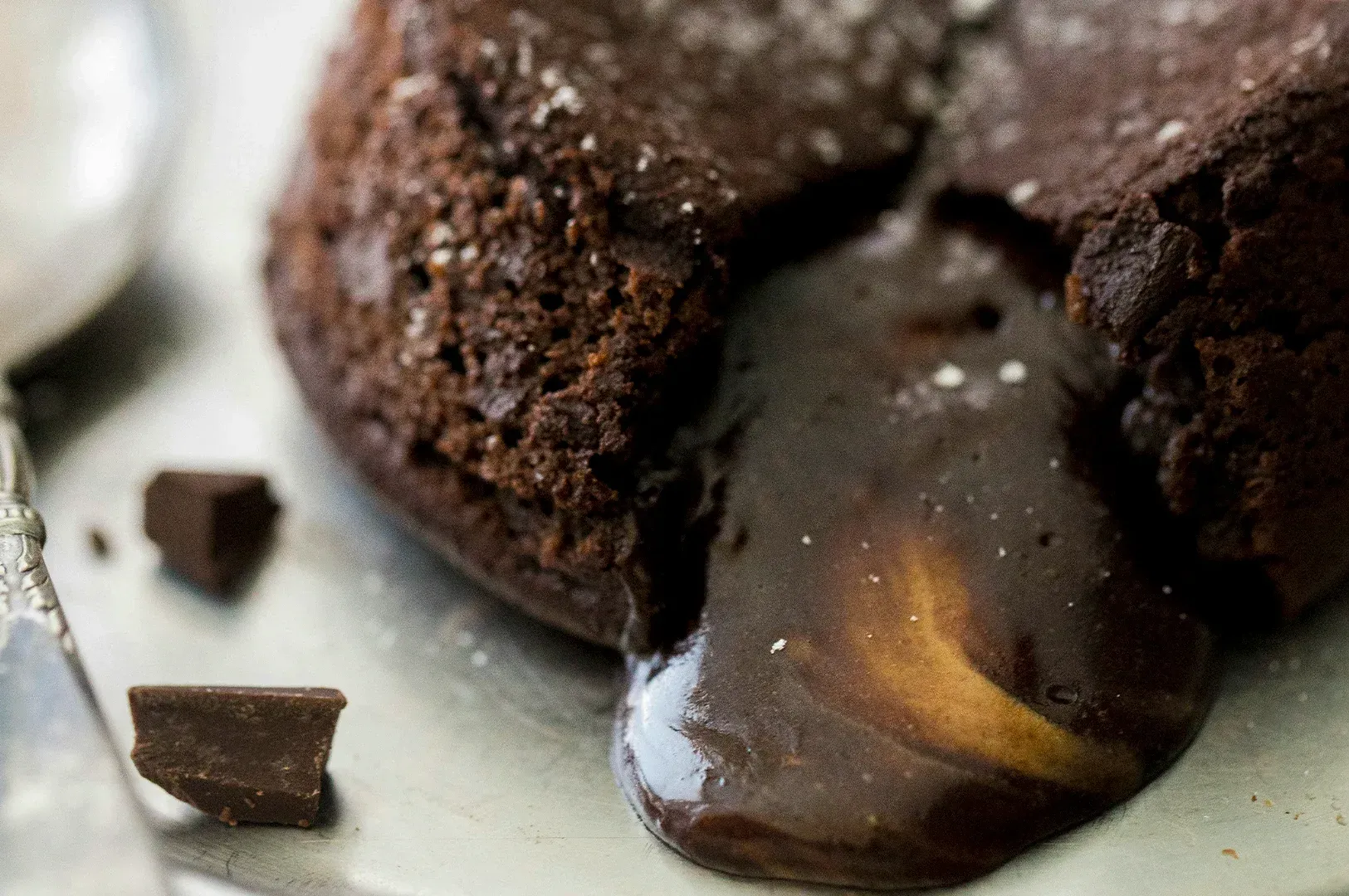 Warm chocolate lava cake that has been sliced open, revealing a thick, glossy, and indulgent dark chocolate center flowing onto a matte silver plate. The cake itself has a rustic, textured crust with a slightly aerated, sponge-like crumb, dusted lightly with fine granules of sugar or sea salt. In the soft-focus background, a vintage silver spoon rests near a few jagged chunks of dark chocolate, enhancing the rich, decadent atmosphere of the dessert. The lighting is soft and natural, emphasizing the contrast between the matte exterior of the cake and the satiny, liquid sheen of the molten 