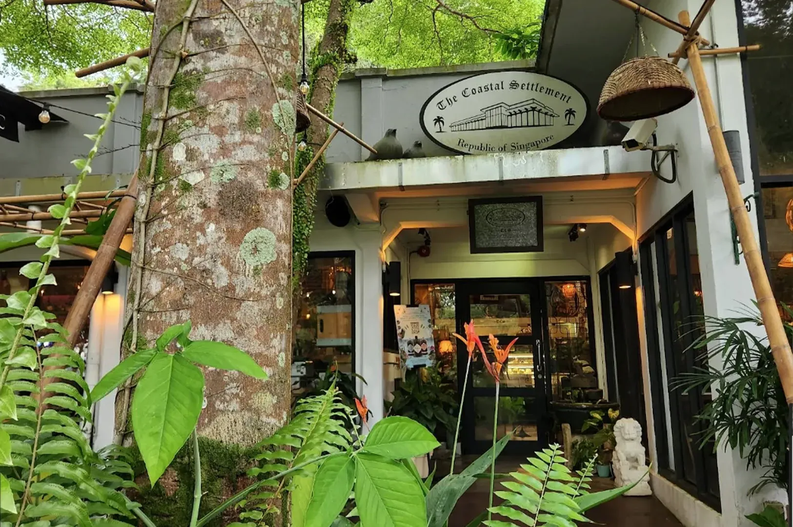 Cozy café entrance with lush greenery, warm lights, and a hanging sign under trees