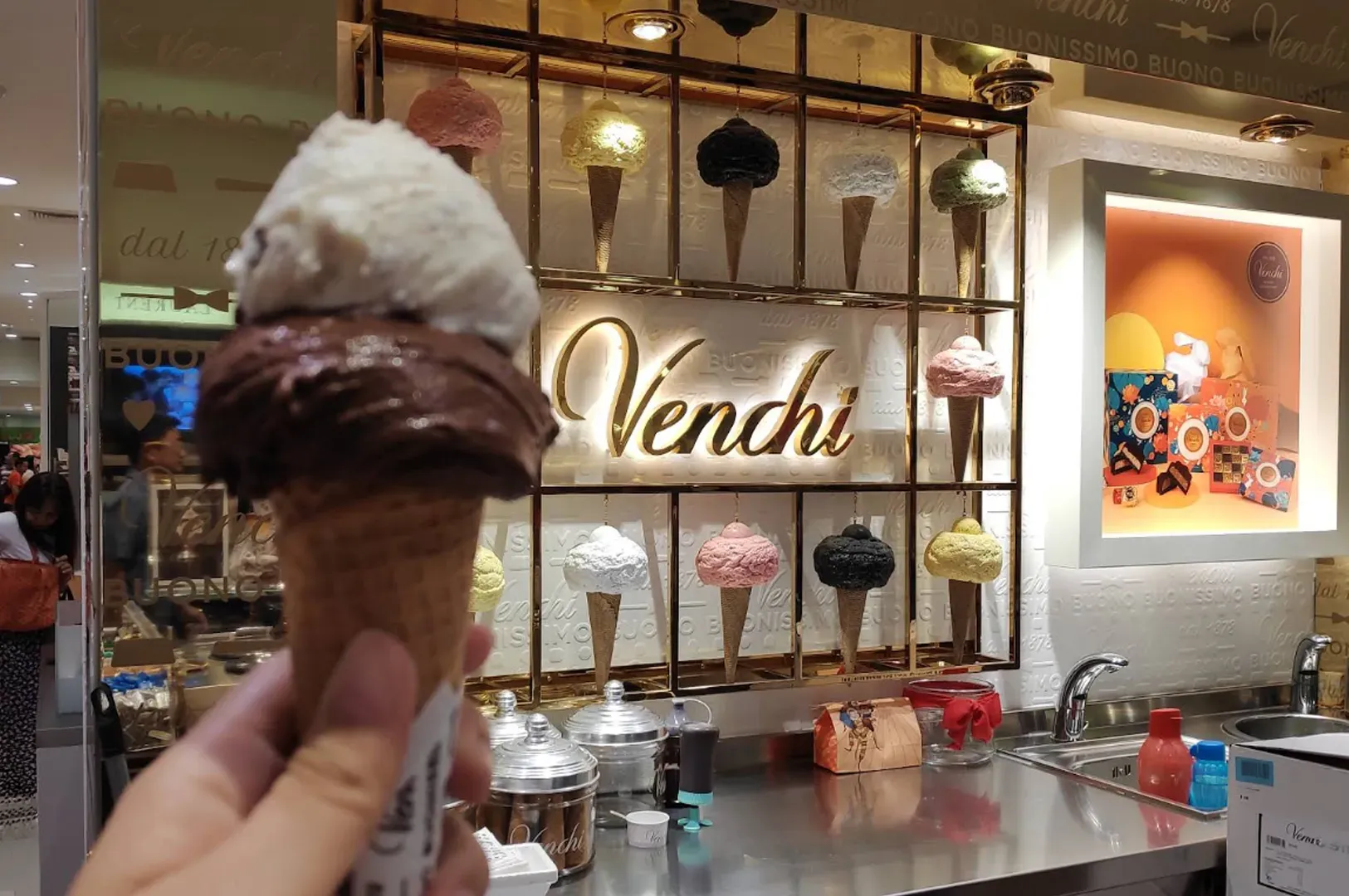 Hand holding a chocolate and vanilla gelato cone in front of a Venchi ice cream shop counter