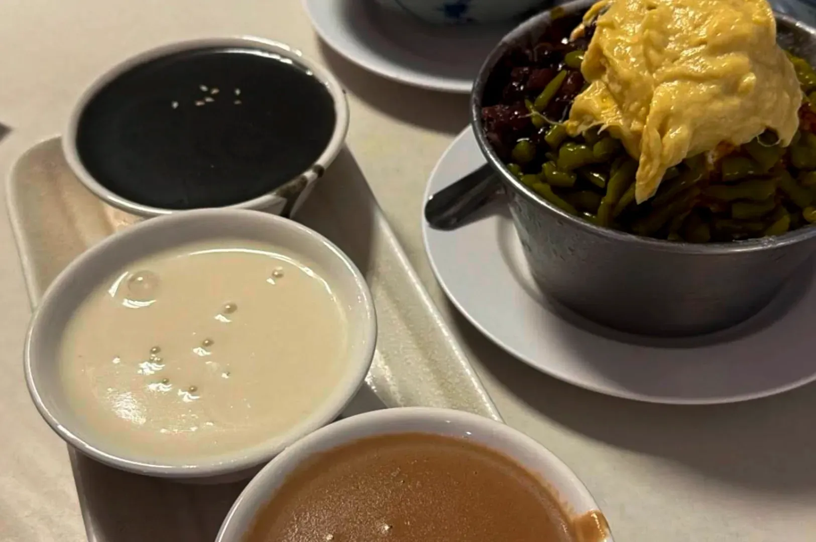 A metal bowl of beans topped with egg next to three small bowls of black, white, and brown dessert soups on a tray.