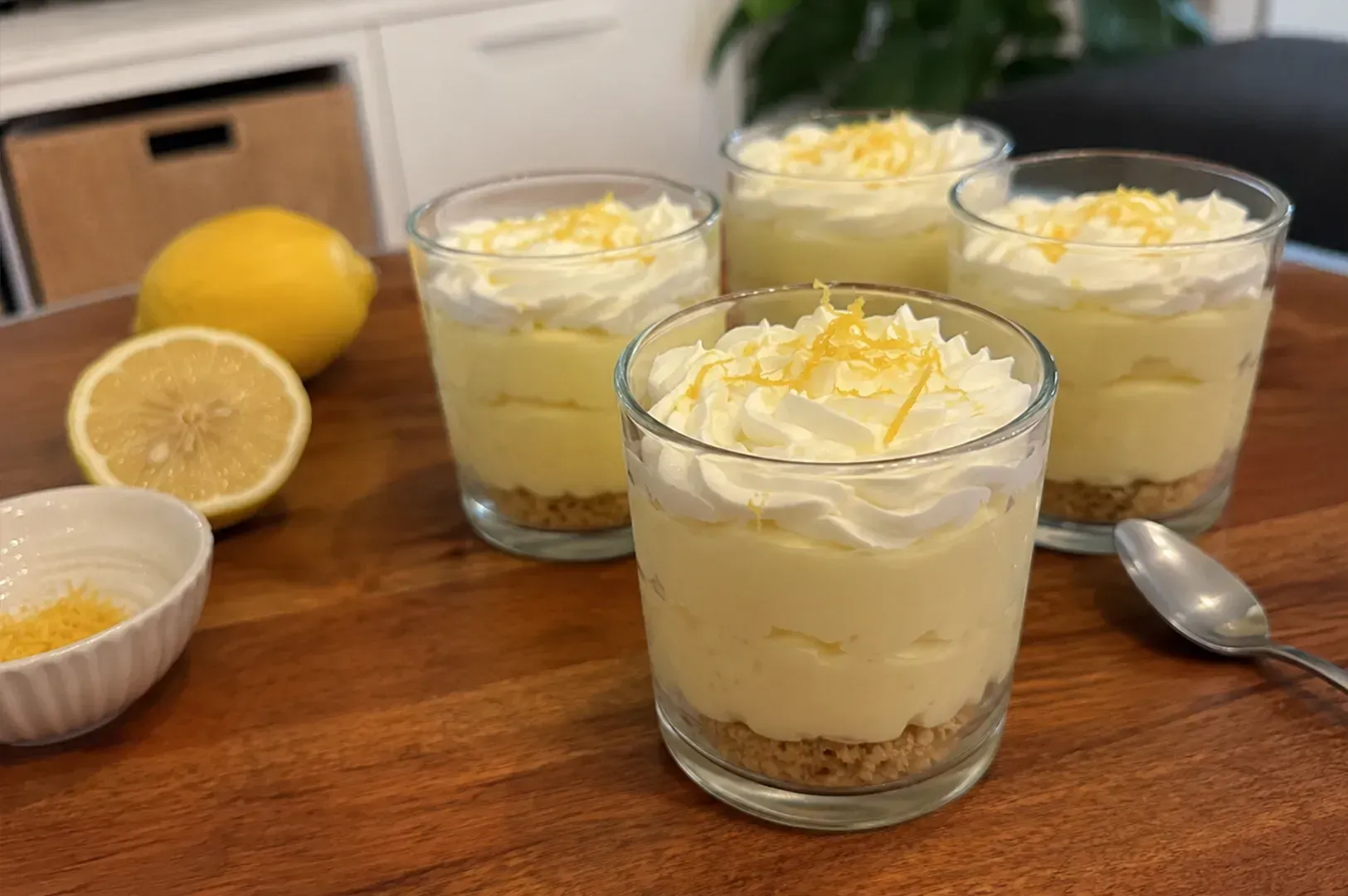 Lemon dessert cups topped with whipped cream and zest on a wooden table, with fresh lemons nearby