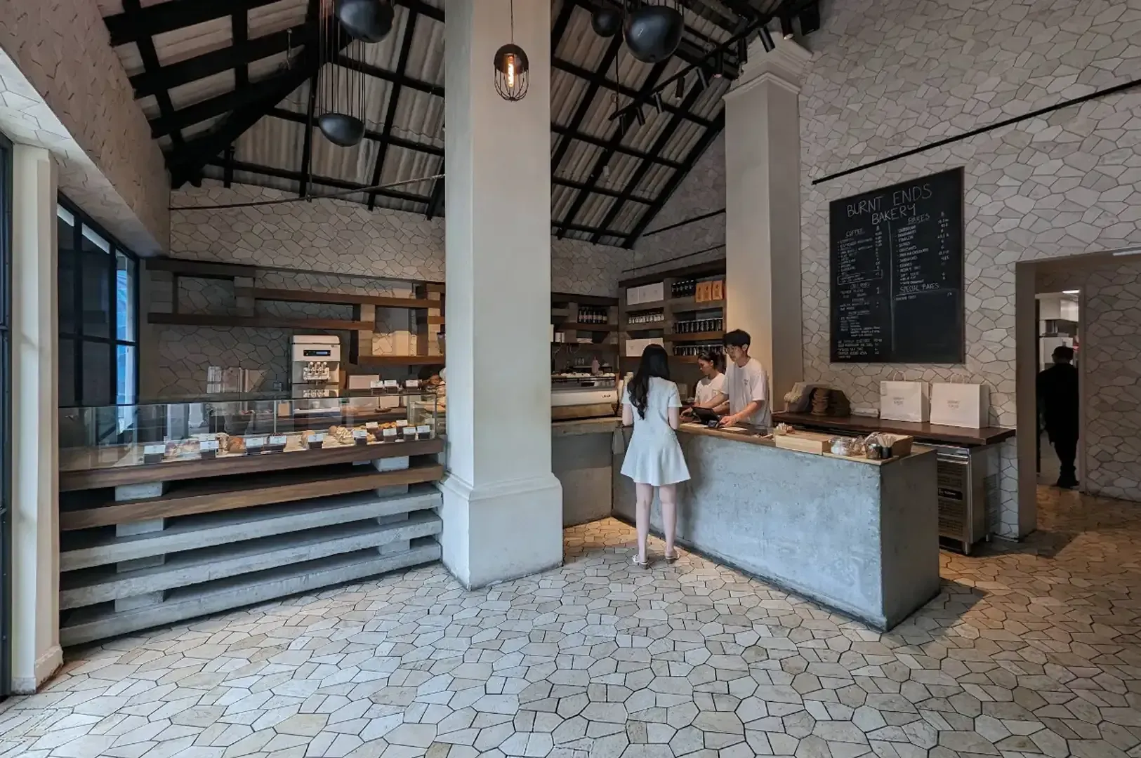 Modern café interior with display counter, high ceilings, and two people standing at the counter.