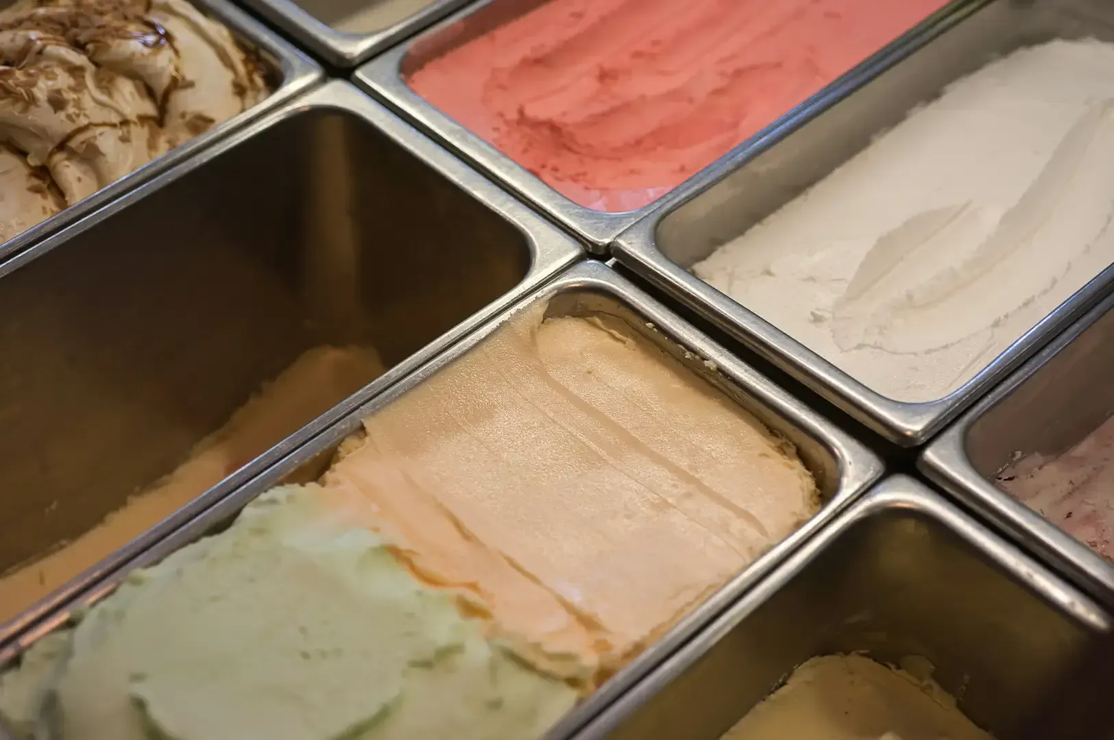 Assorted gelato flavors in metal trays, including pink, white, green, and tan scoops.