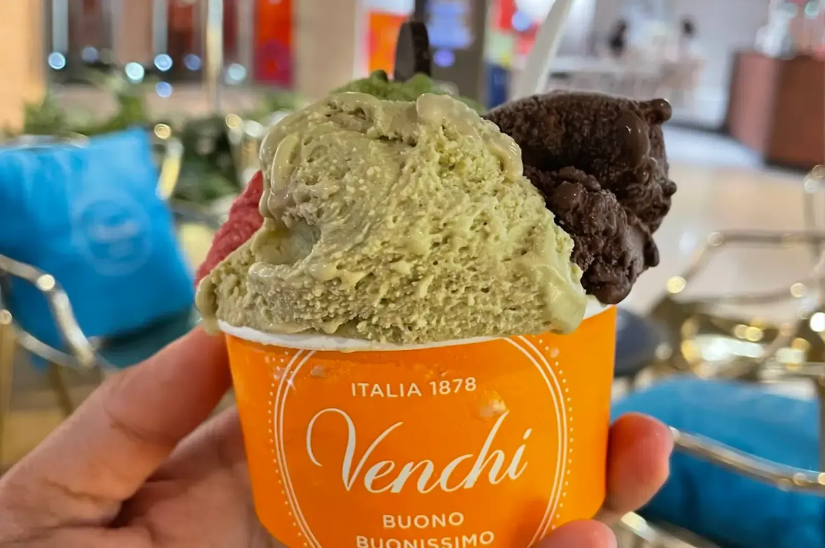 Hand holding a Venchi gelato cup with green and dark scoops, blurred café lights in background