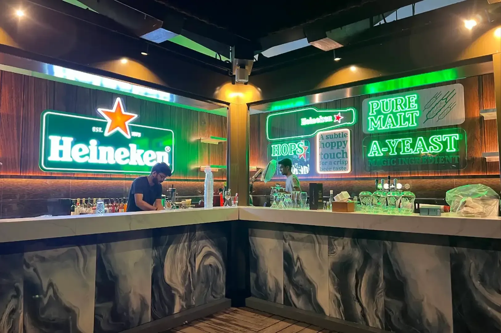 Neon-lit bar counter with Heineken signs and green “Drink until at least” wall slogan.