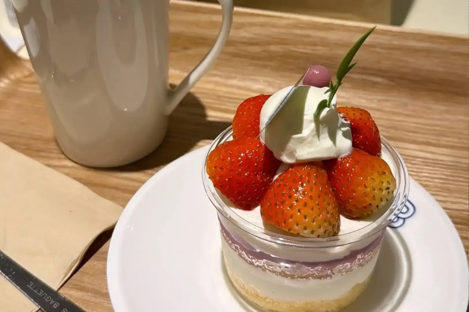 Strawberry shortcake topped with whipped cream on a white plate beside a mug on a wooden table