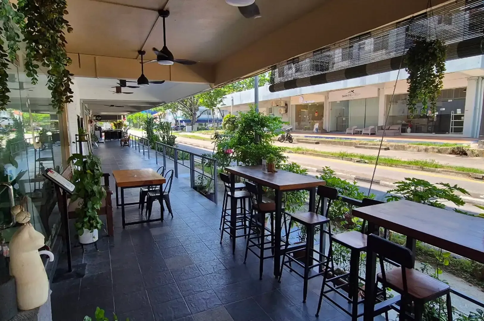 Open-air café seating along a street, with tables, stools, and hanging plants under a covered awning.