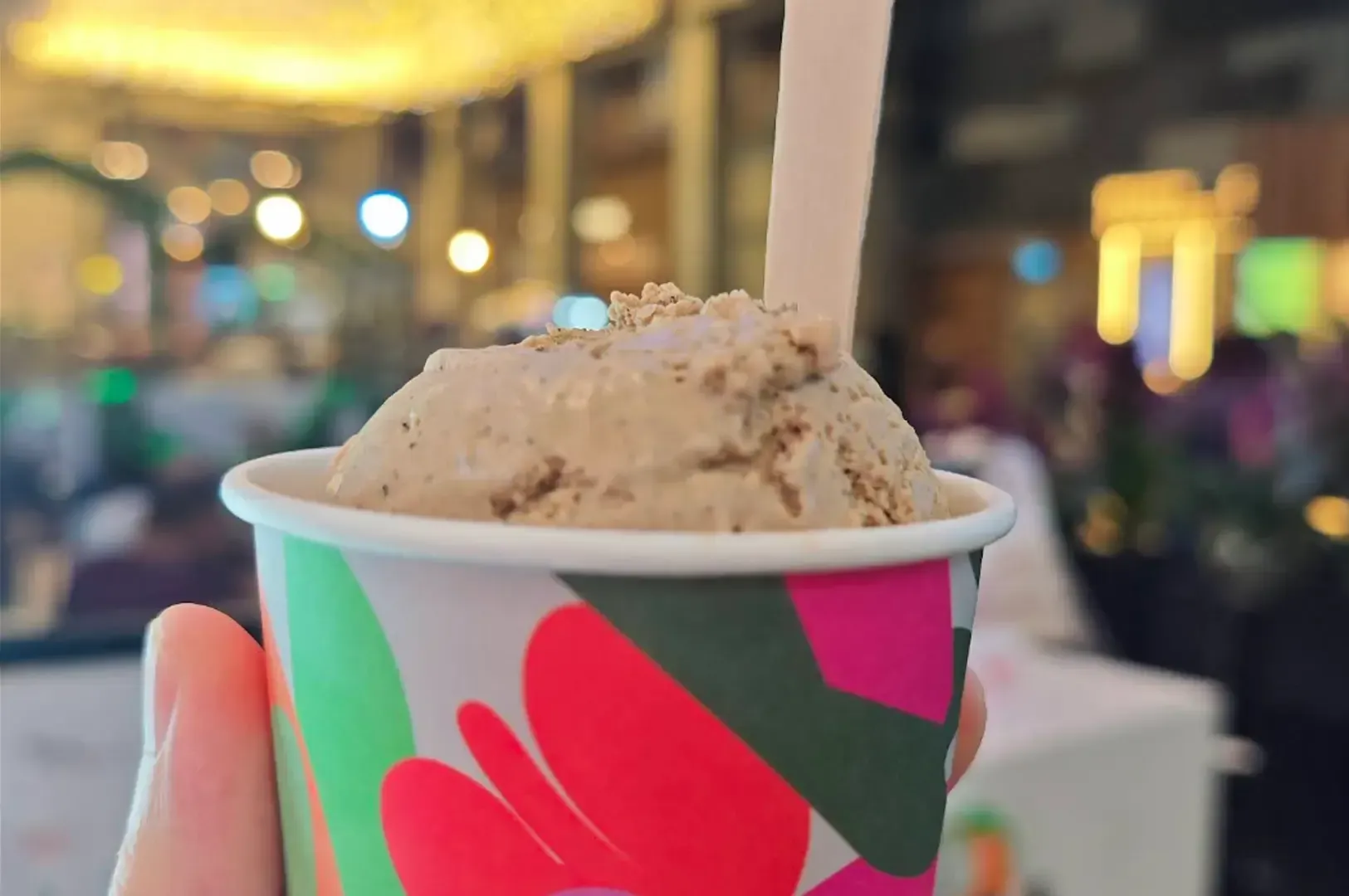 Cup of ice cream with a straw in a colorful paper cup, held up in a blurred indoor café setting