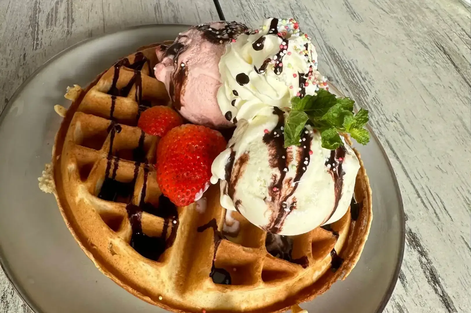 Waffle topped with strawberries, whipped cream, chocolate drizzle, and mint on a gray plate