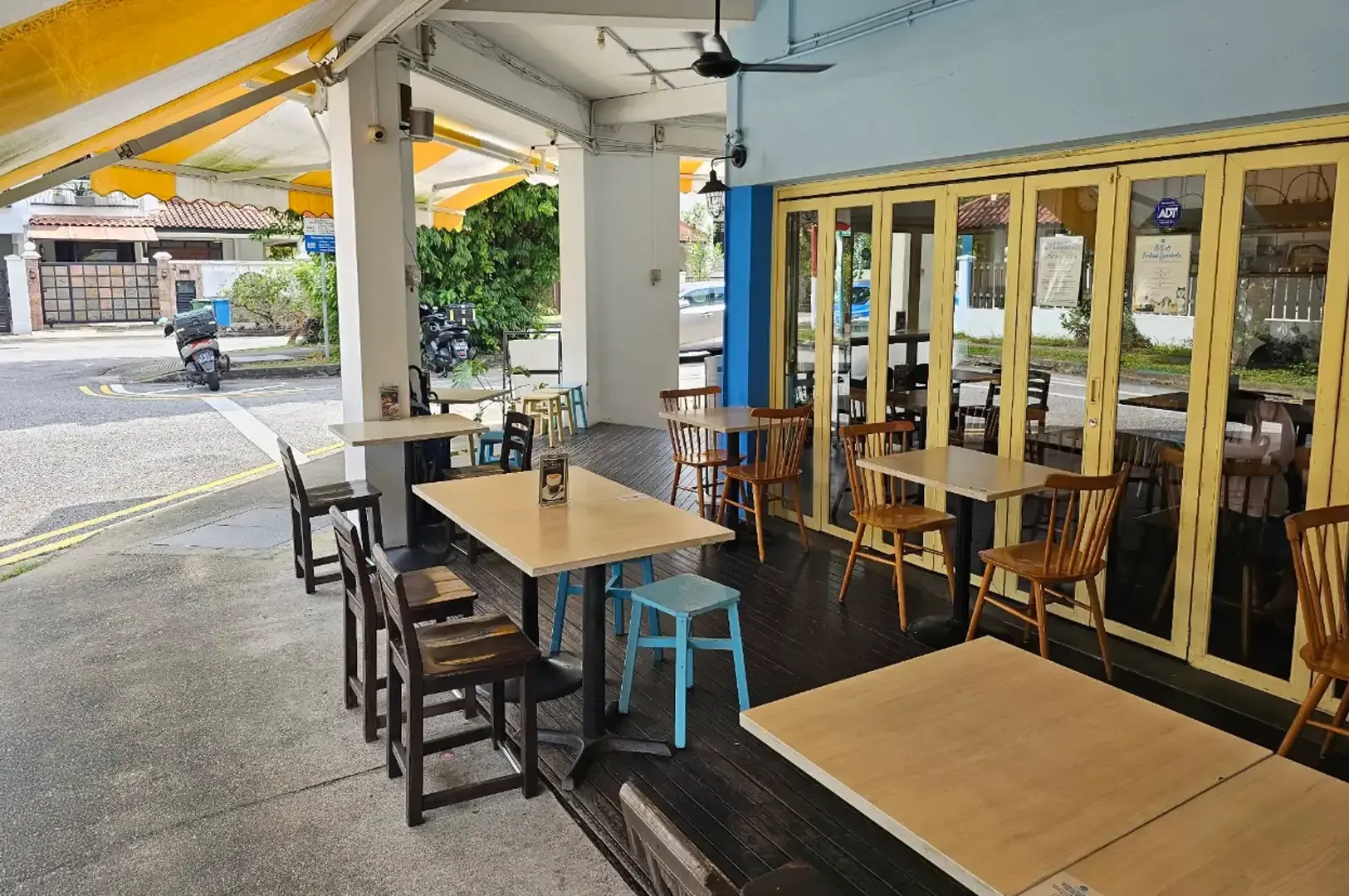 Outdoor café seating with wooden tables and stools under a covered walkway, beside large glass doors.