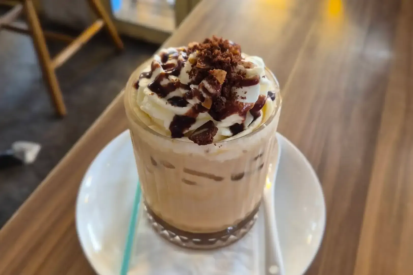 Whipped cream-topped iced coffee in a glass on a saucer with a straw on a wooden table