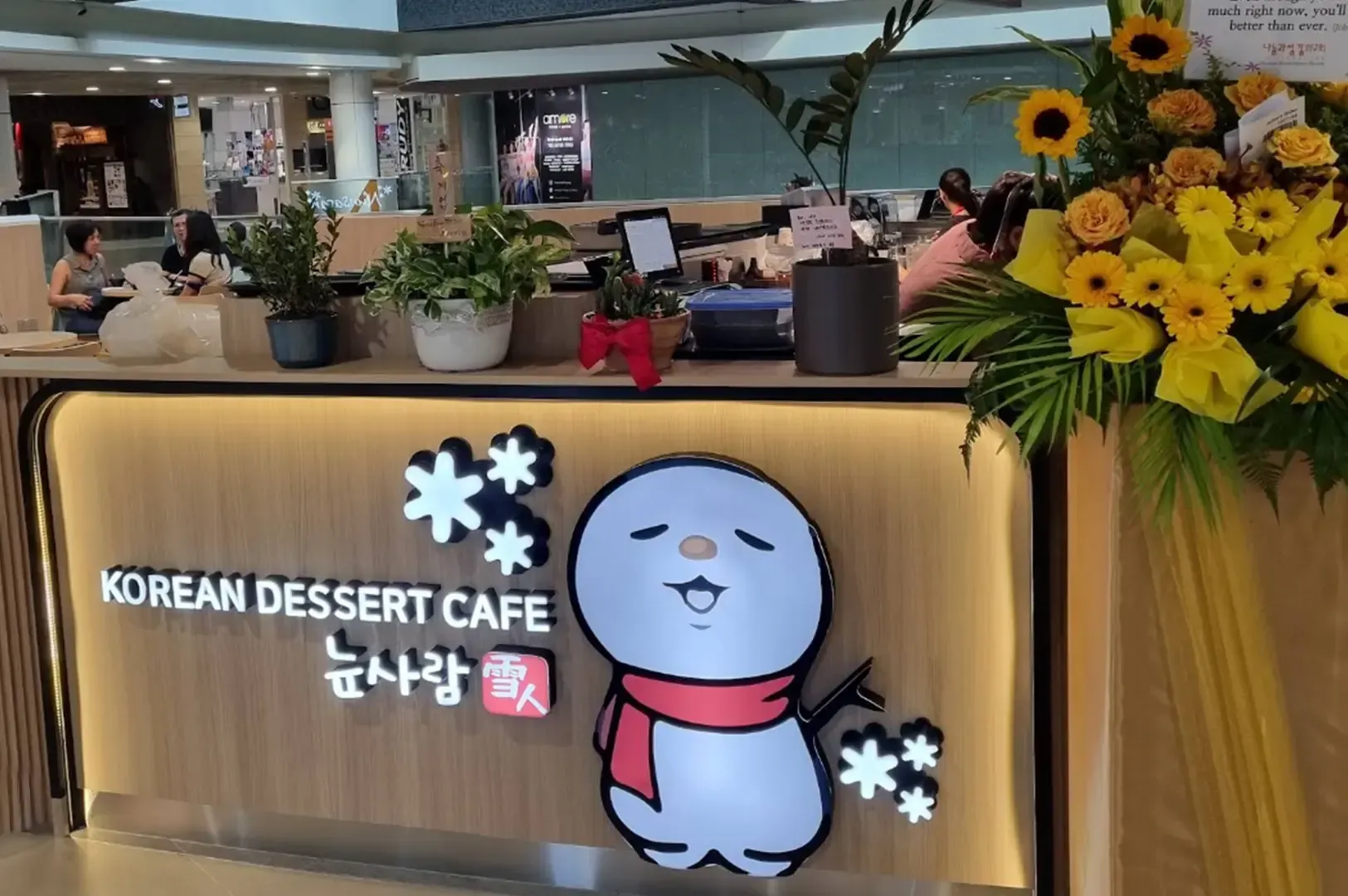 Korean dessert cafe counter with cartoon mascot sign, warm wood paneling, and yellow flowers
