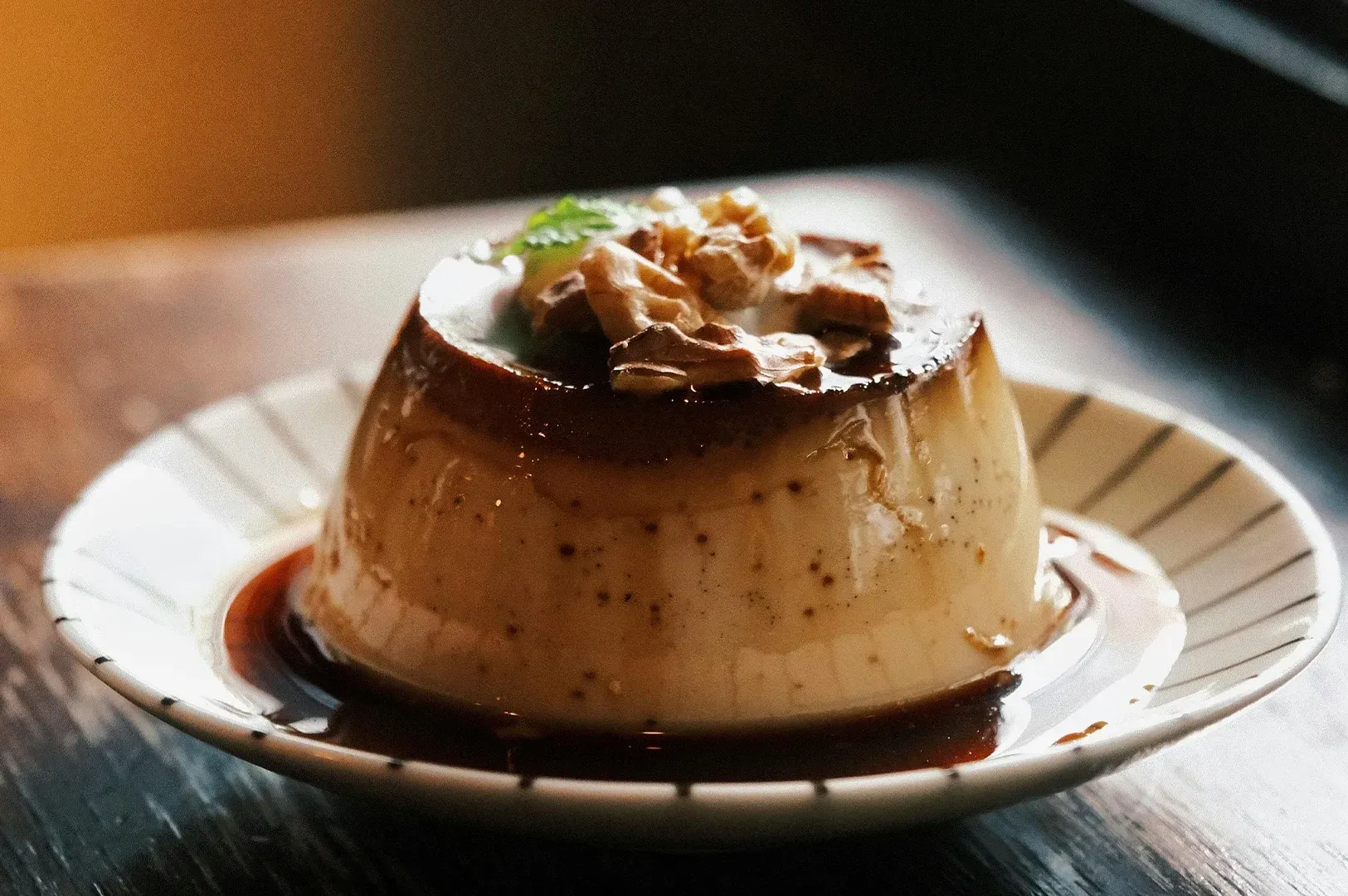 Caramel flan topped with whipped cream on a white plate, drizzled with sauce on a wooden table