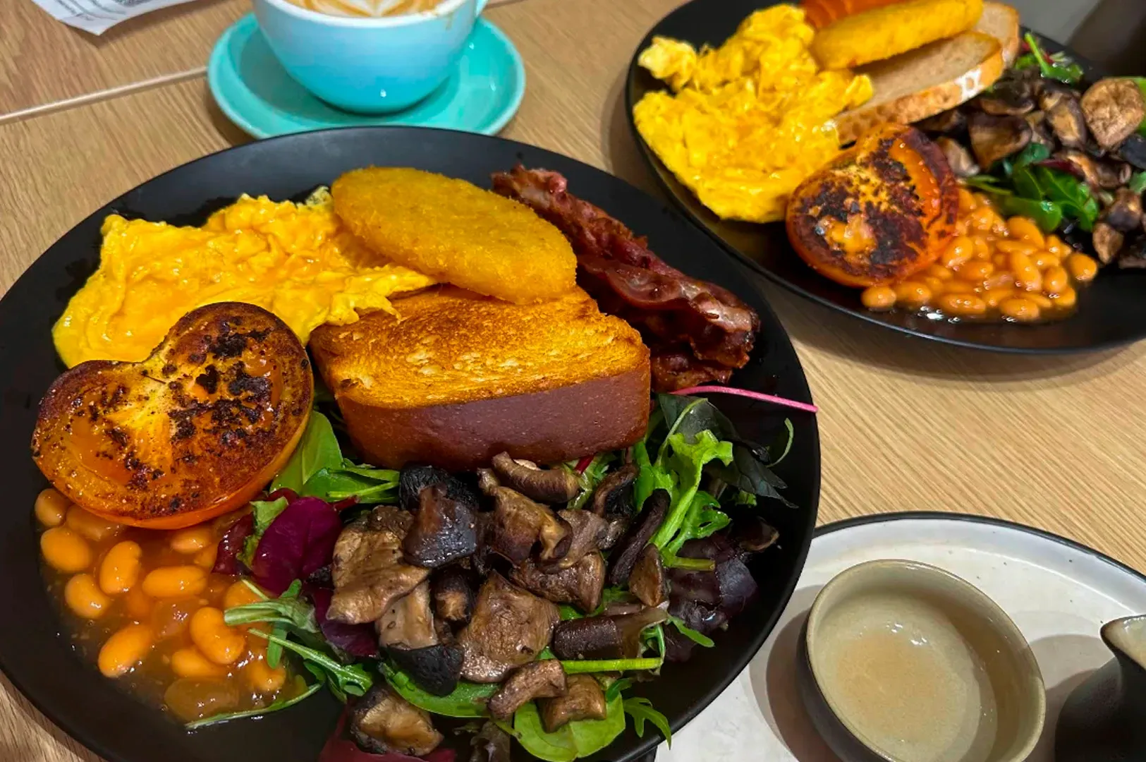 A top-down close-up of two hearty breakfast plates served on a light wooden table. Each black plate is piled with a