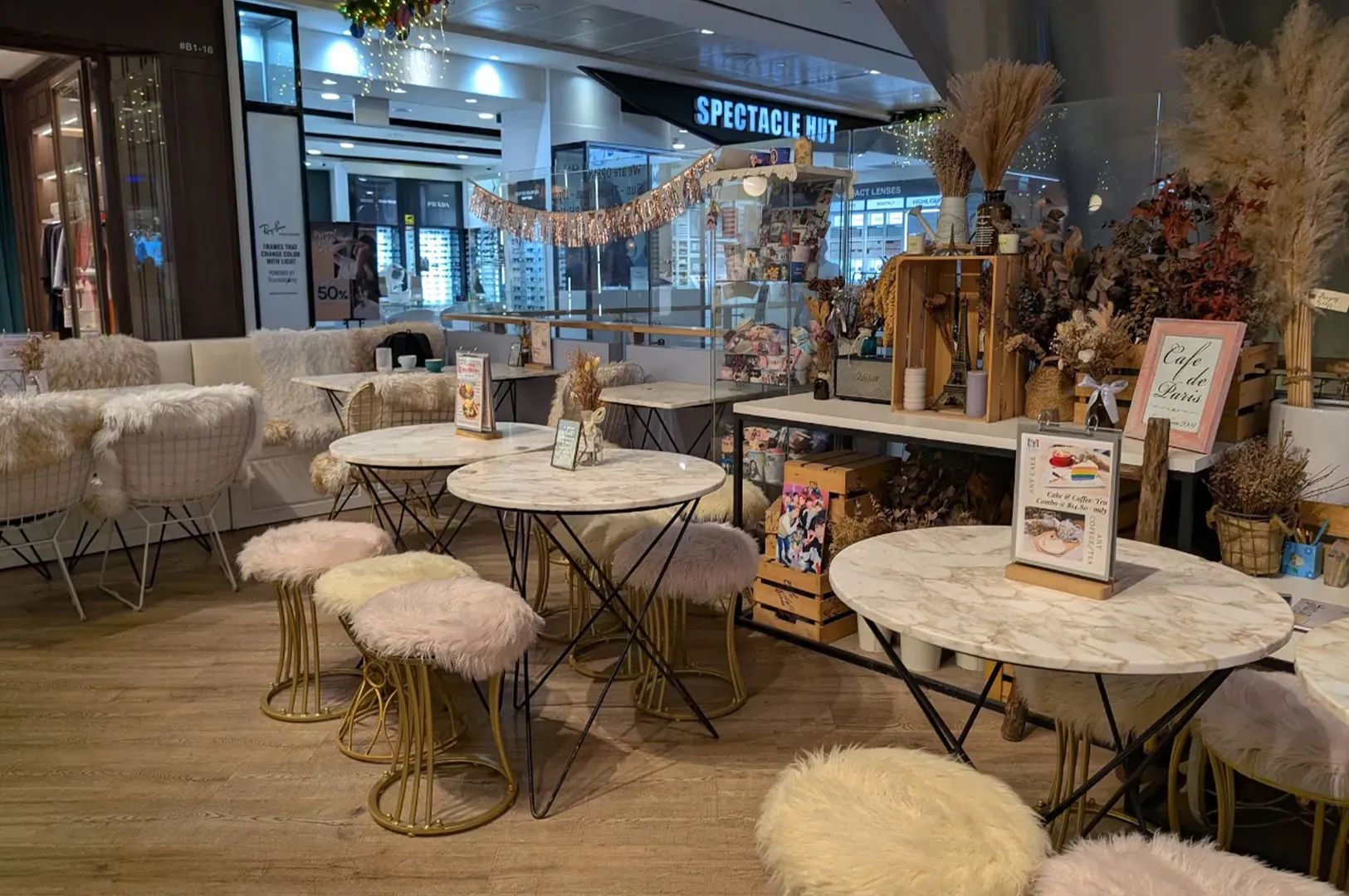 Café seating area with round marble tables, fluffy stools, and dried floral decor inside a mall storefront.