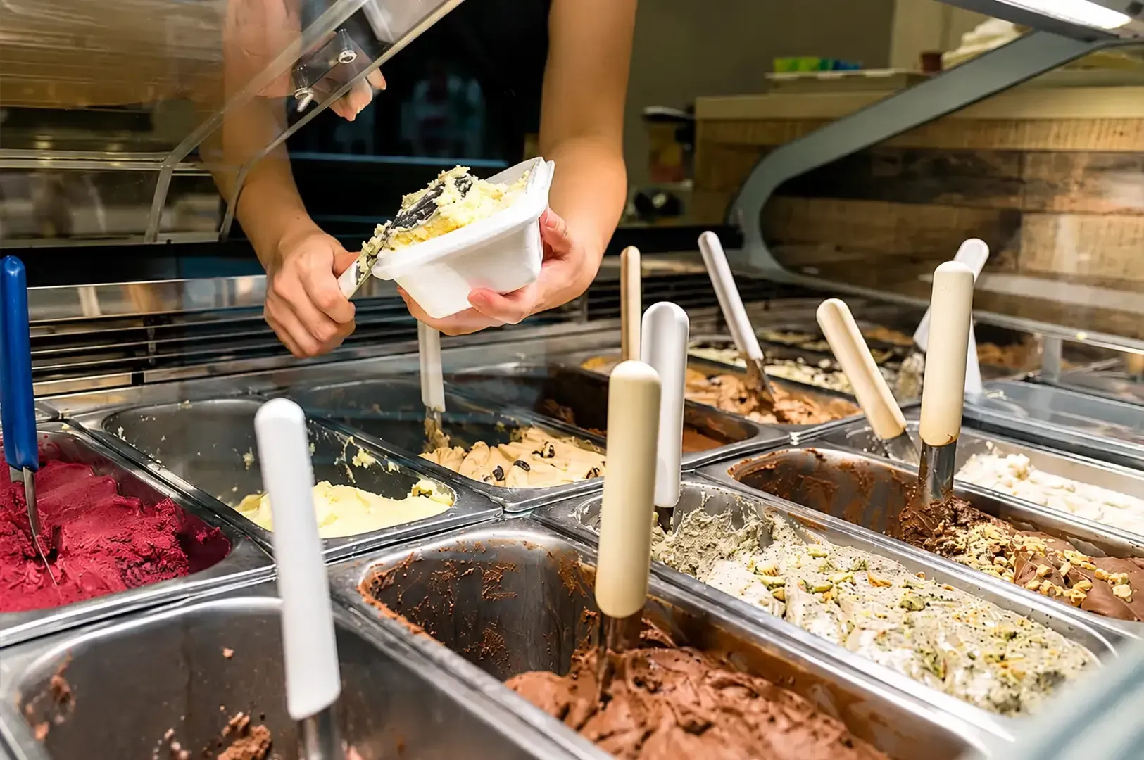 Person serving gelato from a display case with multiple flavors and white scoops.