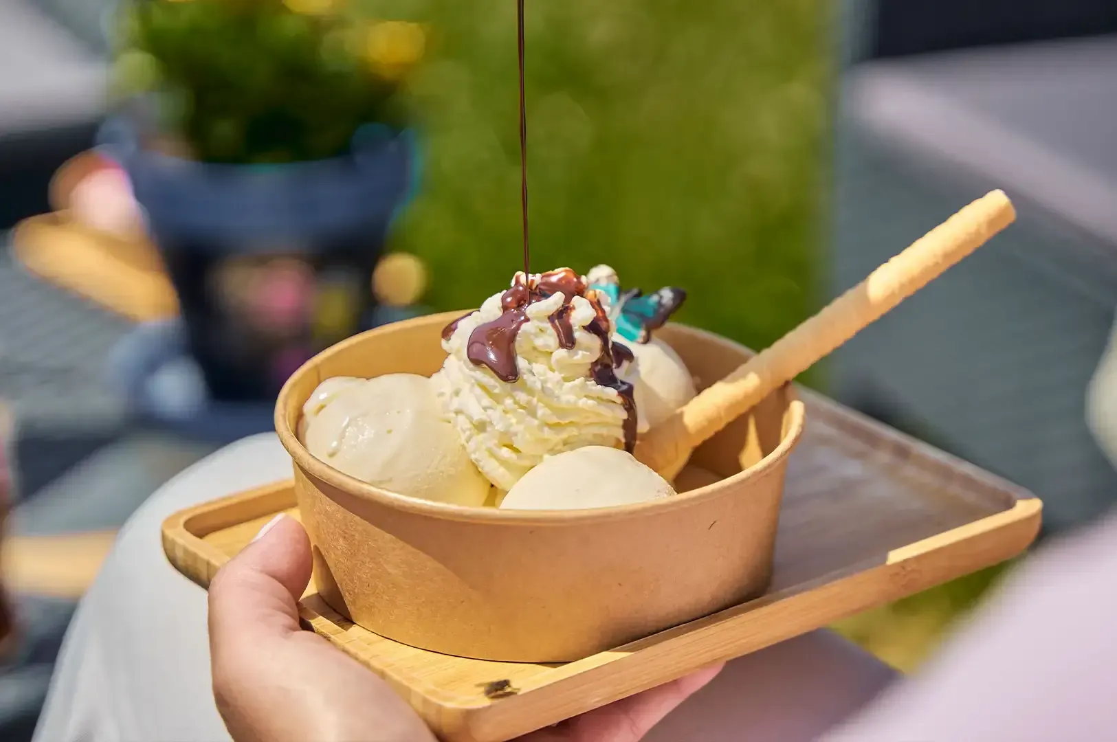 Hand holding a bowl of vanilla ice cream with chocolate sauce and a wafer stick on a tray outdoors