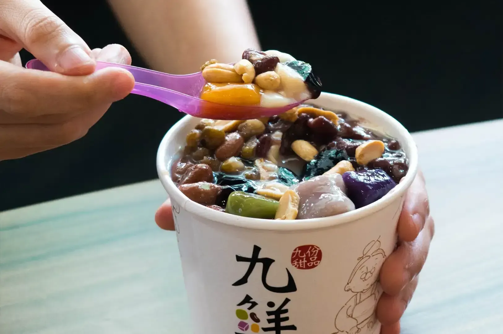 Hand holding a spoon over a cup of colorful shaved ice dessert with toppings and Chinese characters on the cup