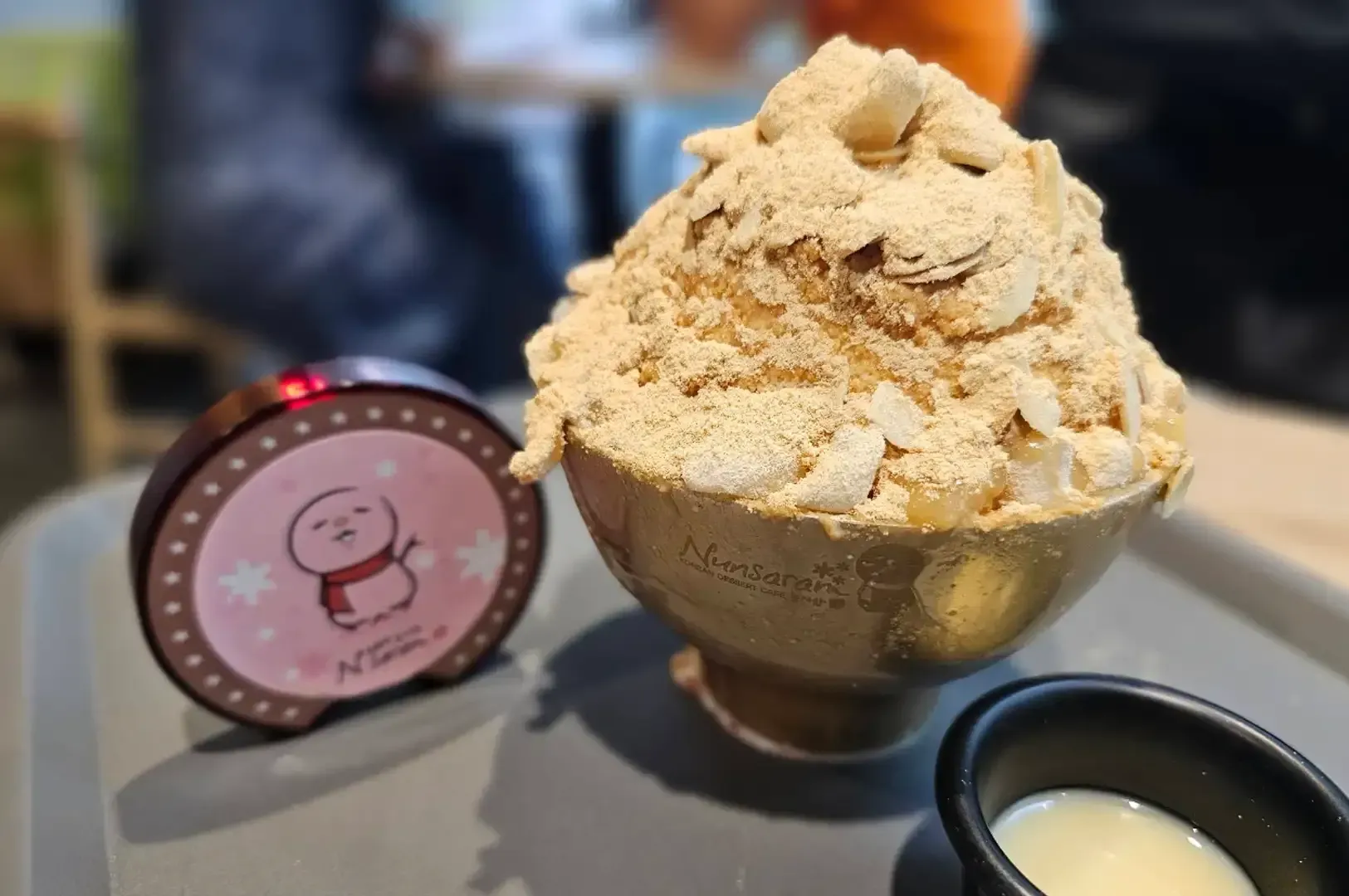 Gold bowl of shaved ice topped with beige powder, beside a round pink tin and a small black cup on a tray.