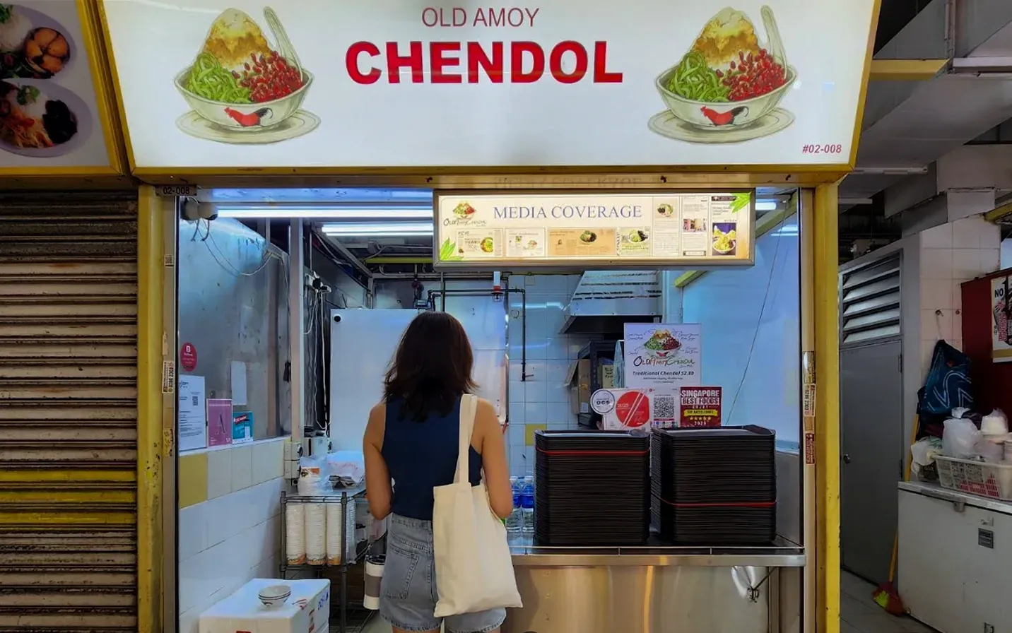 Woman standing at a Chendol dessert stall with a lit sign and counter display