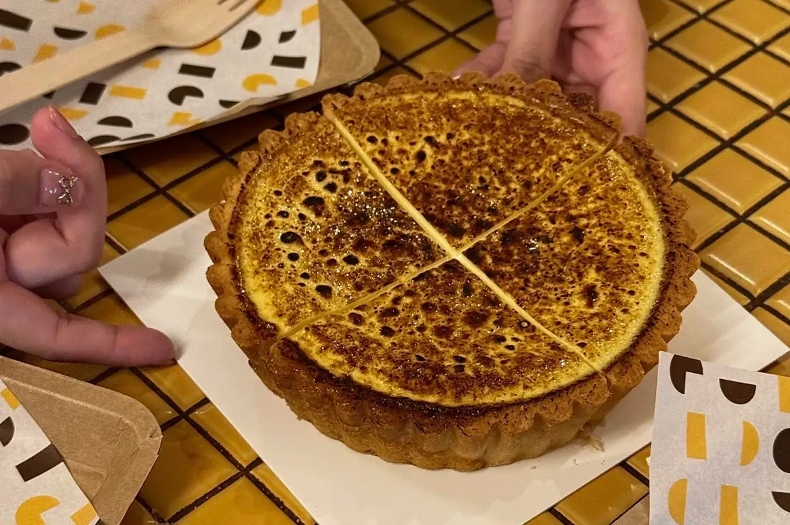 Hands holding a caramelized tart on parchment paper over a checkered tablecloth.