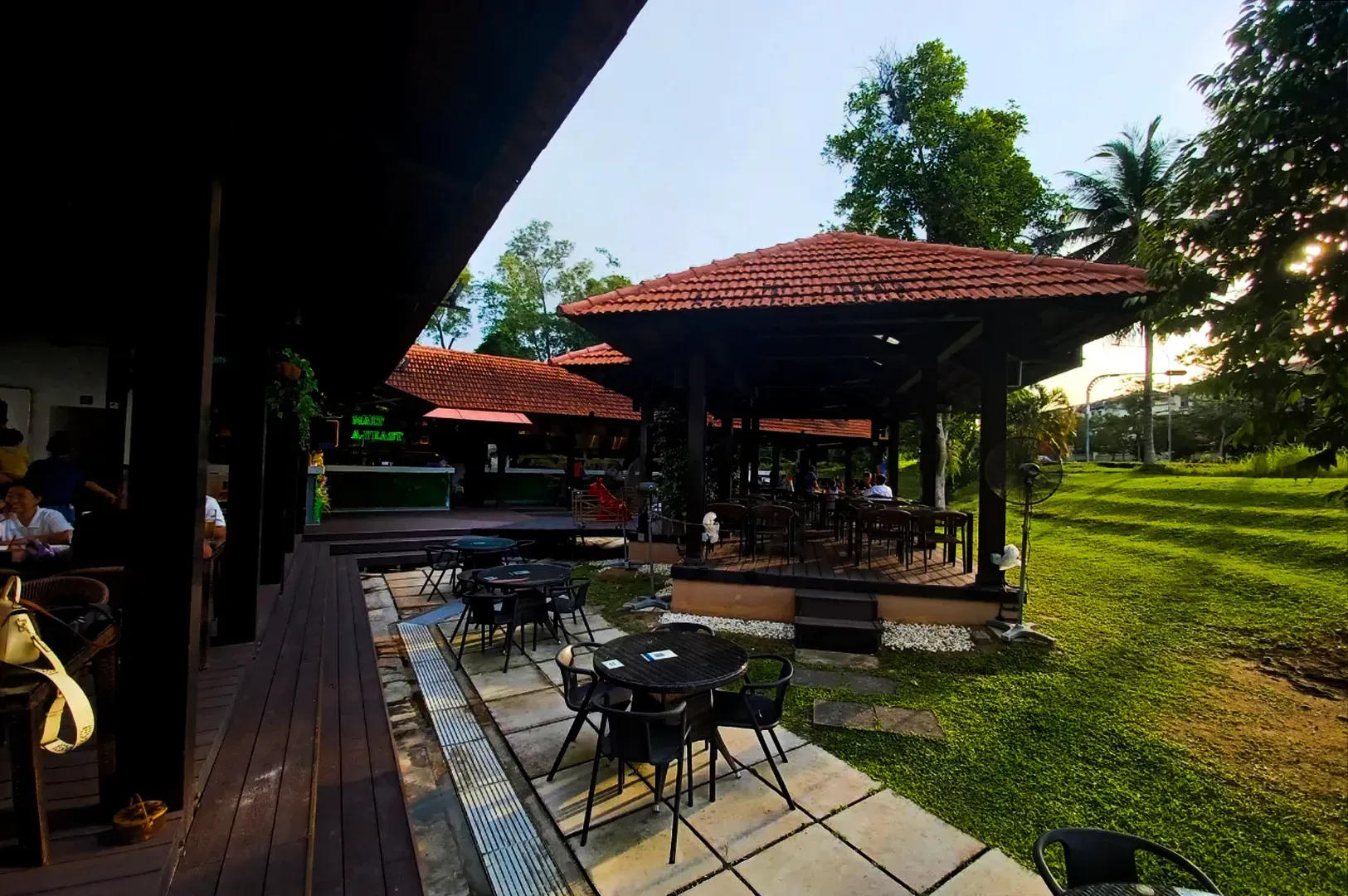 Outdoor café patio with red-tiled umbrellas, tables, and chairs beside a grassy lawn at sunset