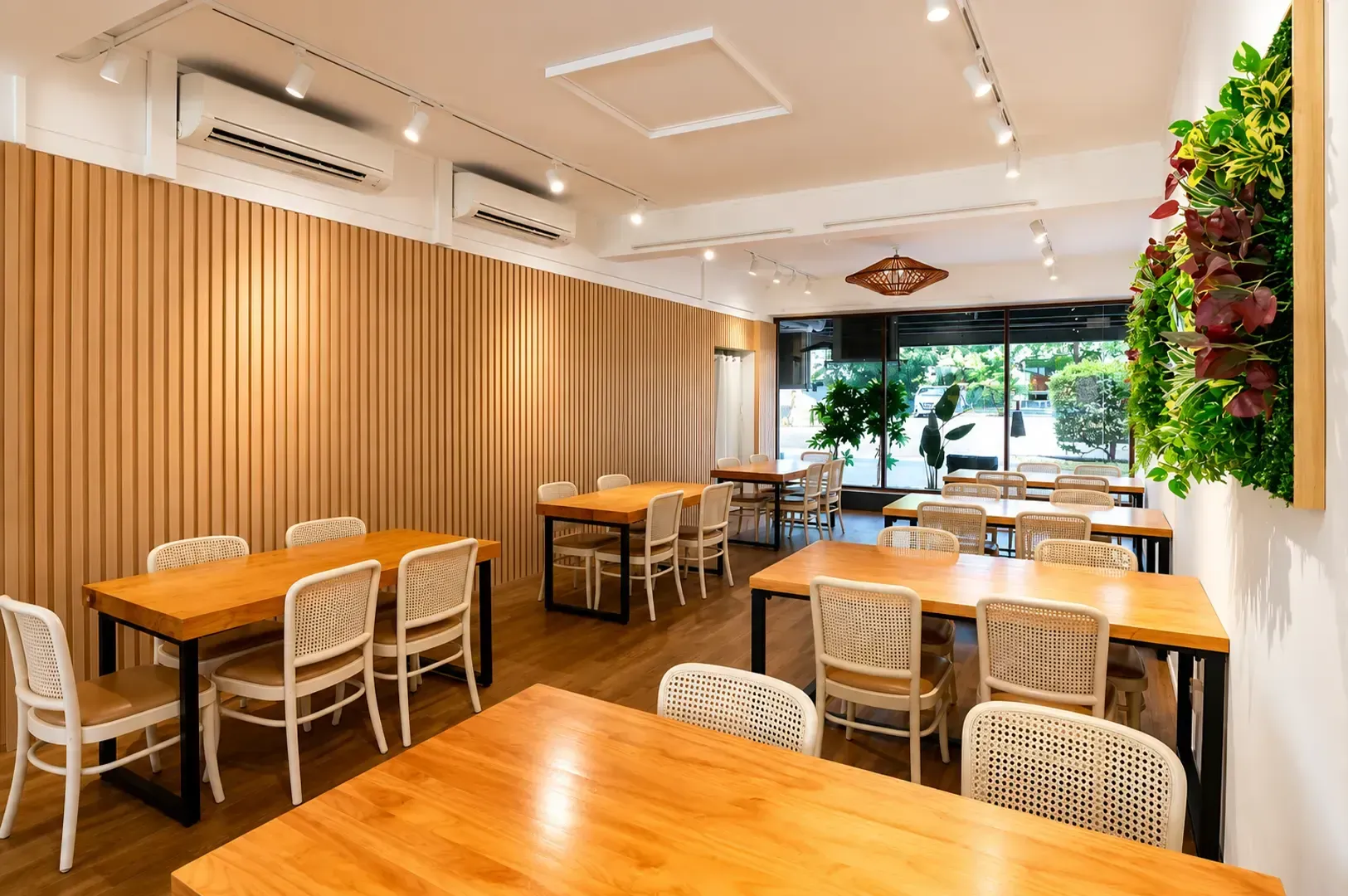 Bright modern cafe dining area with wooden tables, woven chairs, and greenery wall near large windows