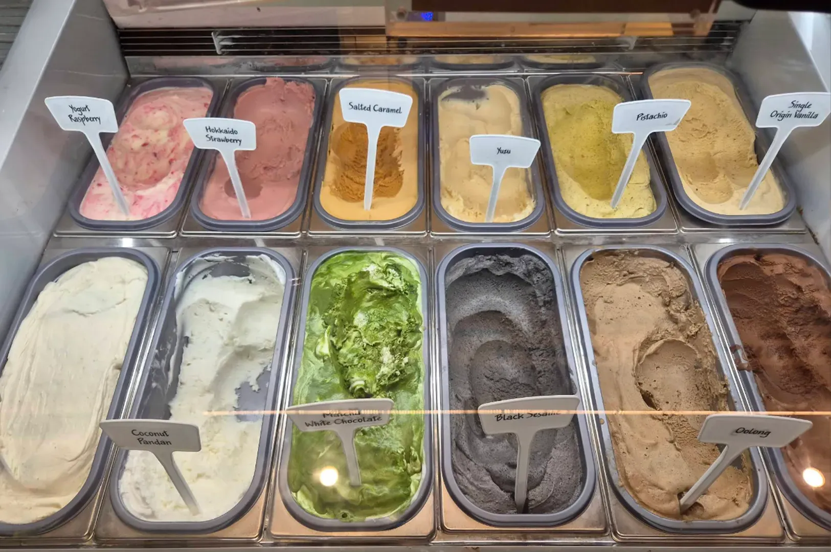 Ice cream display case with assorted flavors in metal tubs, including pink, white, green, dark gray, tan, and brown.