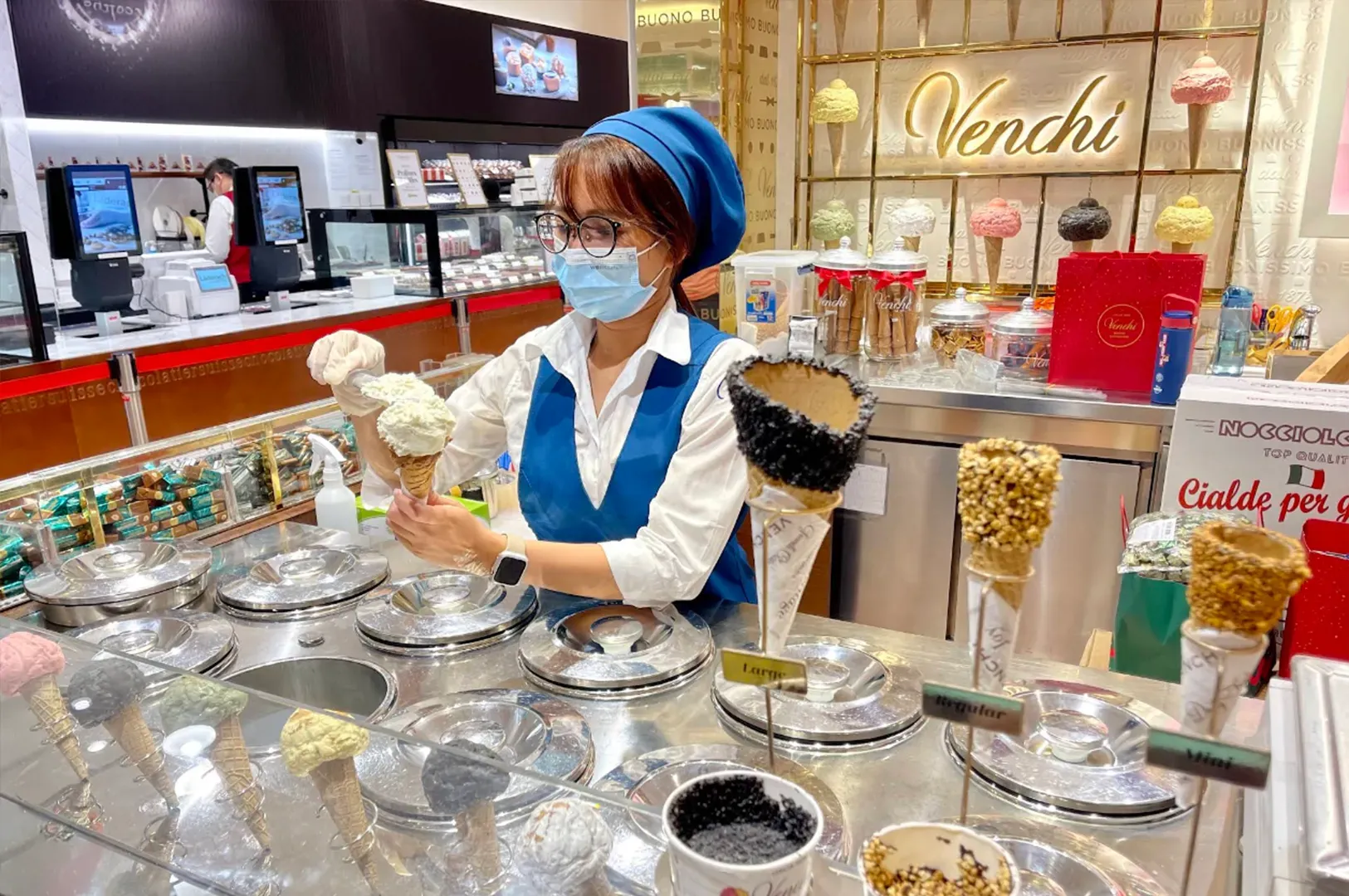 Masked attendant behind gelato counter in bright dessert shop, serving ice cream scoops.