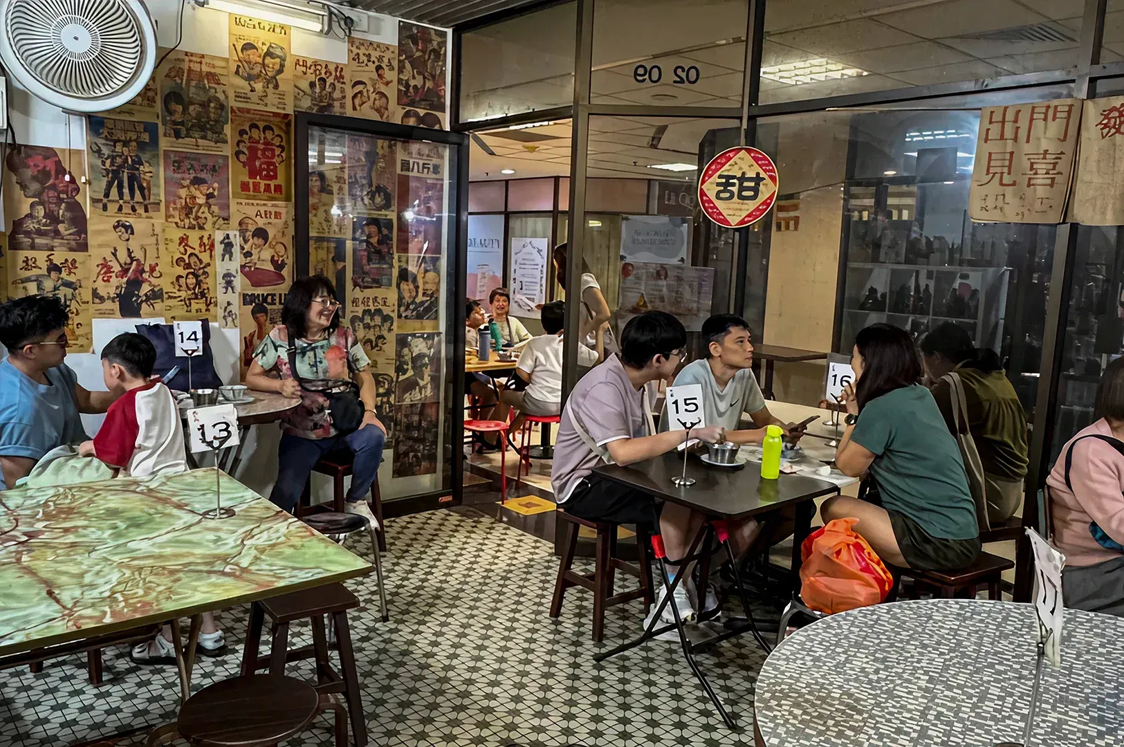 The restaurant's interior exudes a charming, retro vibe, characterized by walls plastered with vintage Chinese movie posters and classic advertisements. The floor is covered in traditional mosaic tiles, and the seating consists of simple wooden stools and small square tables, each marked with a metal number stand. Large glass partitions reveal a glimpse of more seating areas, while a circular red sign with the character for