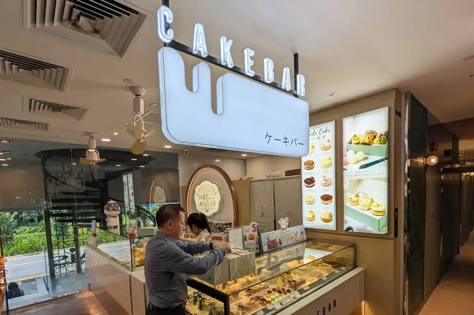 Cakebar dessert shop with display case, lit menu boards, and a person serving behind the counter