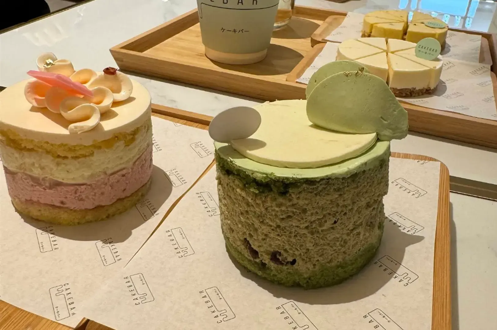 Assorted pastel dessert cakes on a tray, including pink and green mousse cakes with fruit and cream toppings.