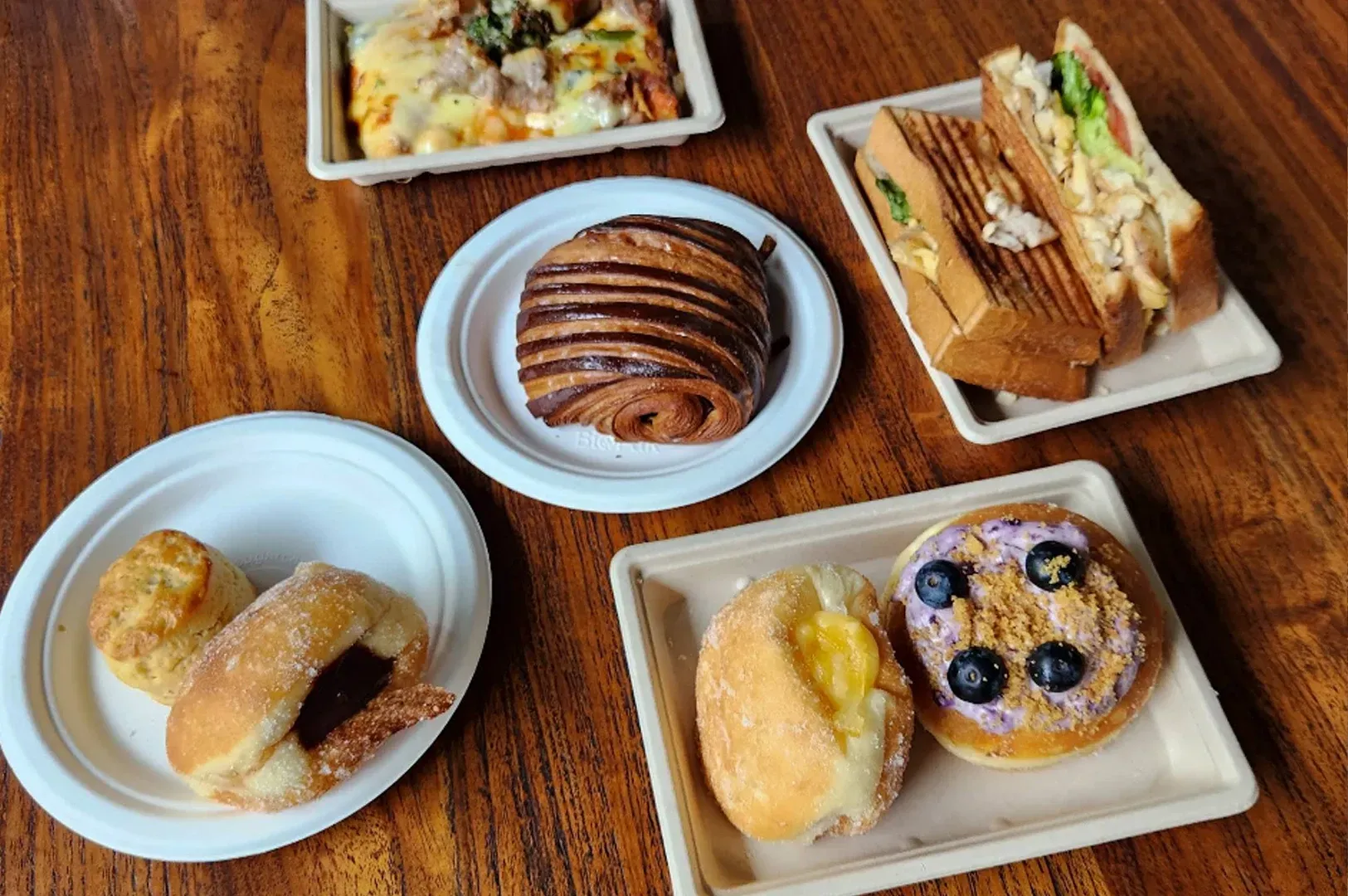 Assorted pastries and sandwiches on small white plates on a wooden table.