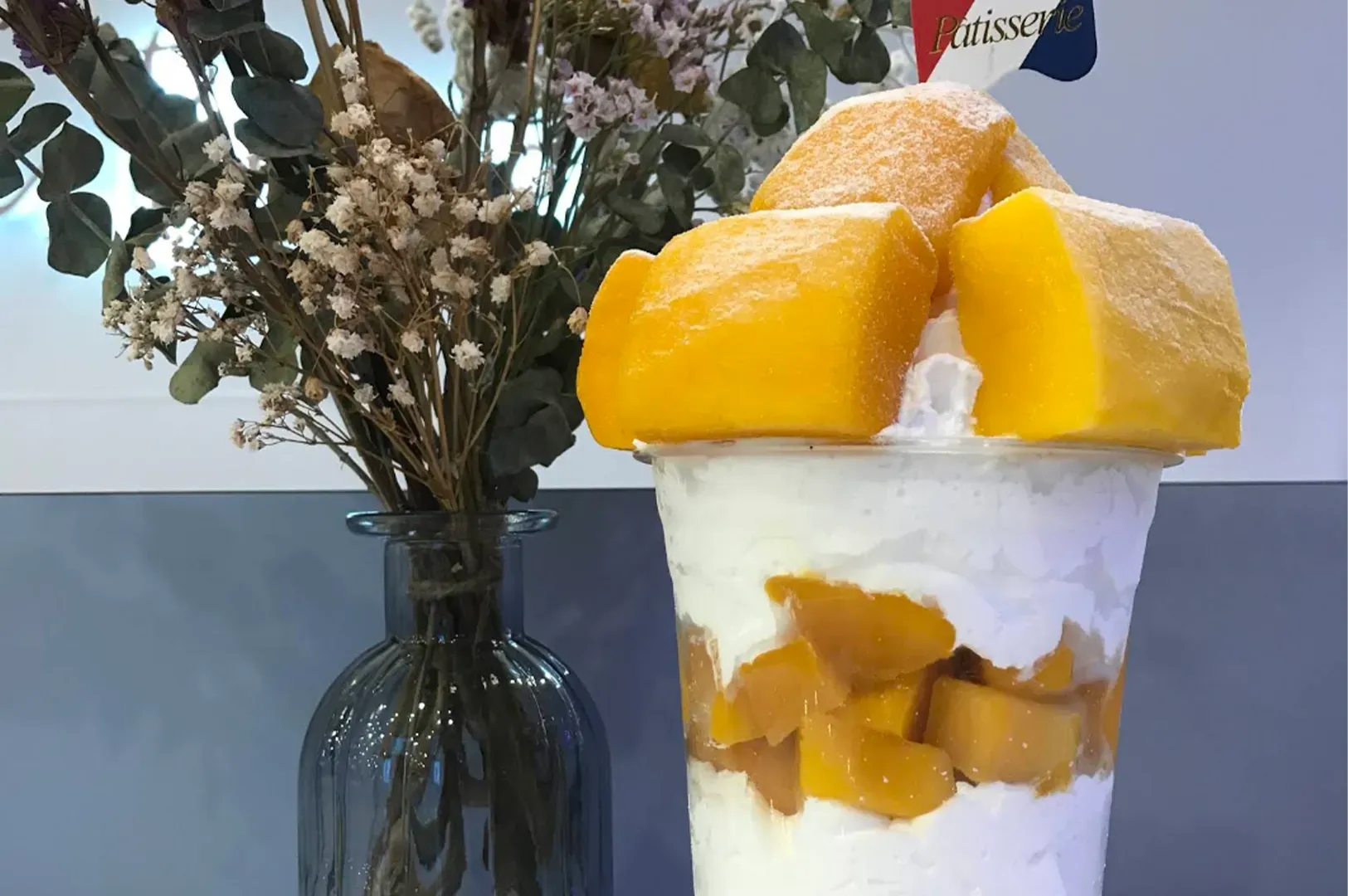 Mango shaved ice in a clear cup topped with fruit chunks, beside a vase of flowers on a table.