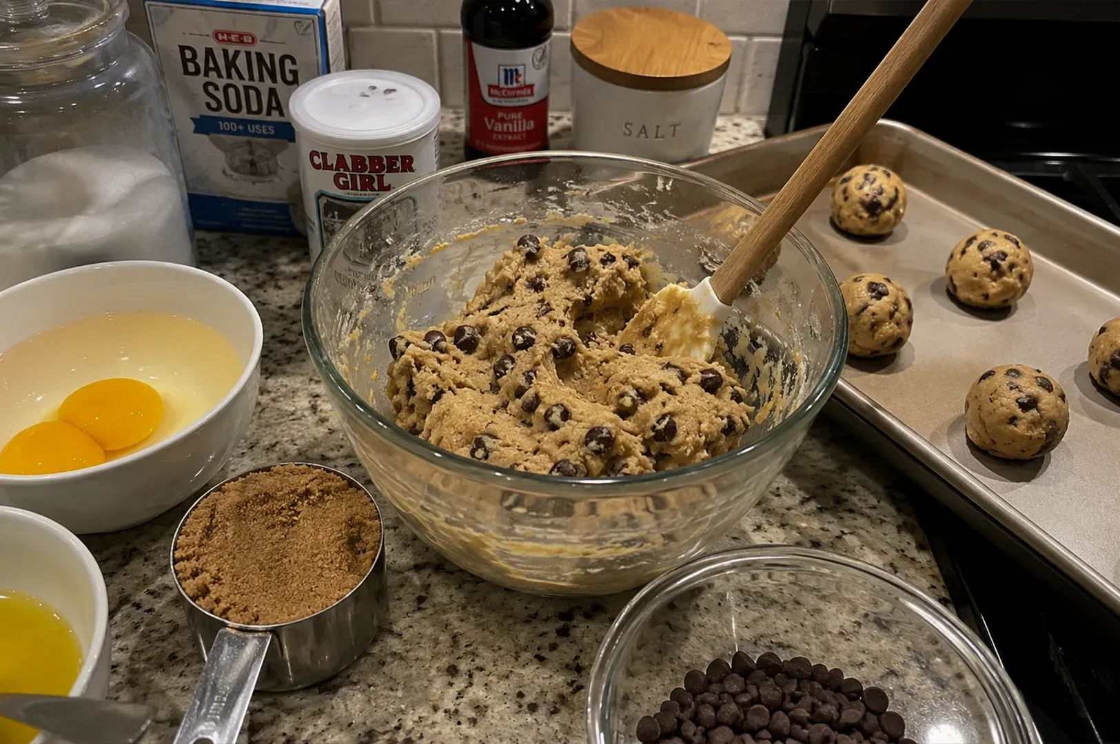 Baking setup with cookie dough bowl, ingredients, and chocolate chip cookie dough balls on trays