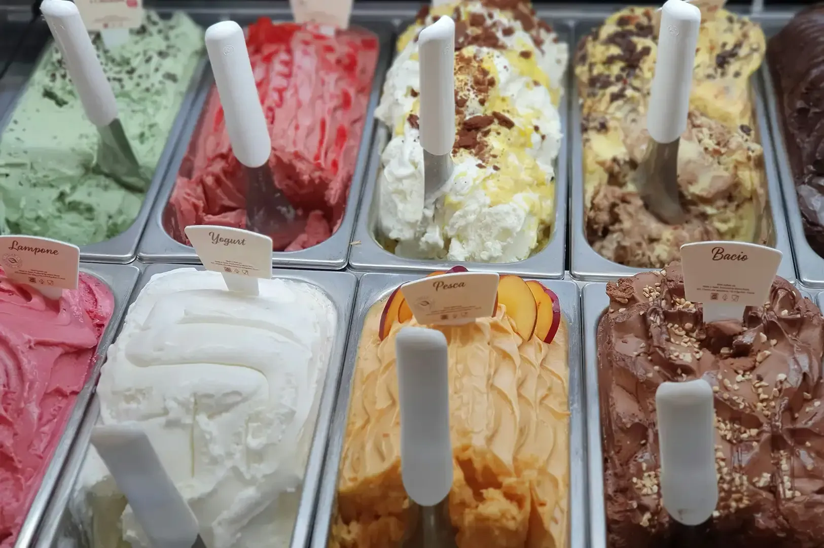 Assorted gelato flavors in display tubs with white serving spoons, including pink, green, white, yellow, and brown.