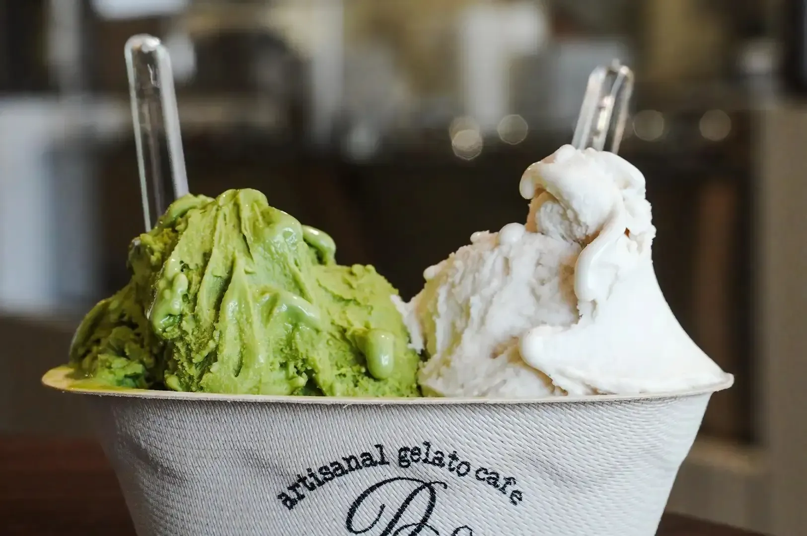 Two scoops of gelato, green and white, in a cup with spoons at an Italian gelato cafe