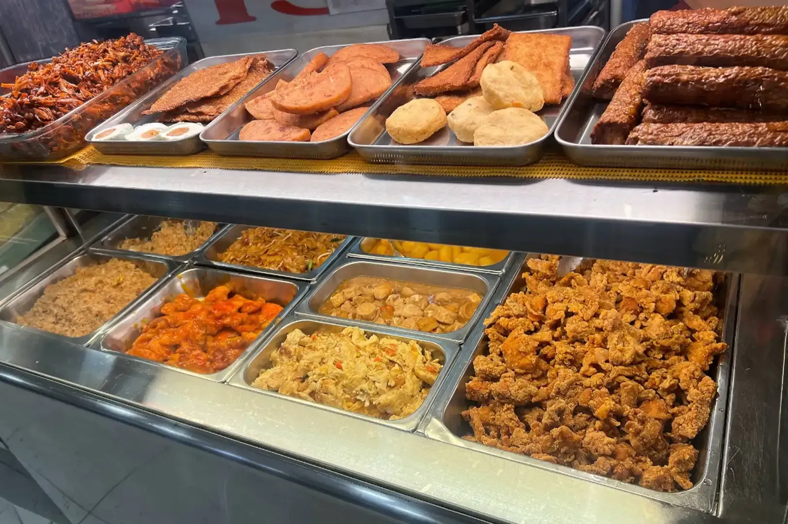 Buffet counter with assorted fried and baked foods in metal trays under glass display