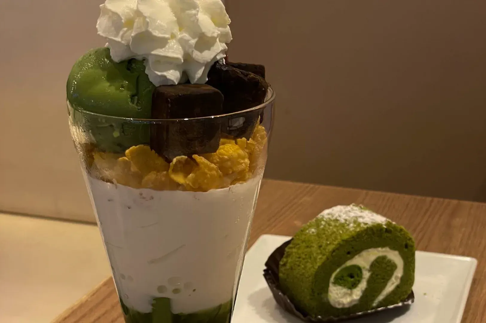 Green dessert parfait topped with whipped cream beside a slice of matcha roll cake on a plate