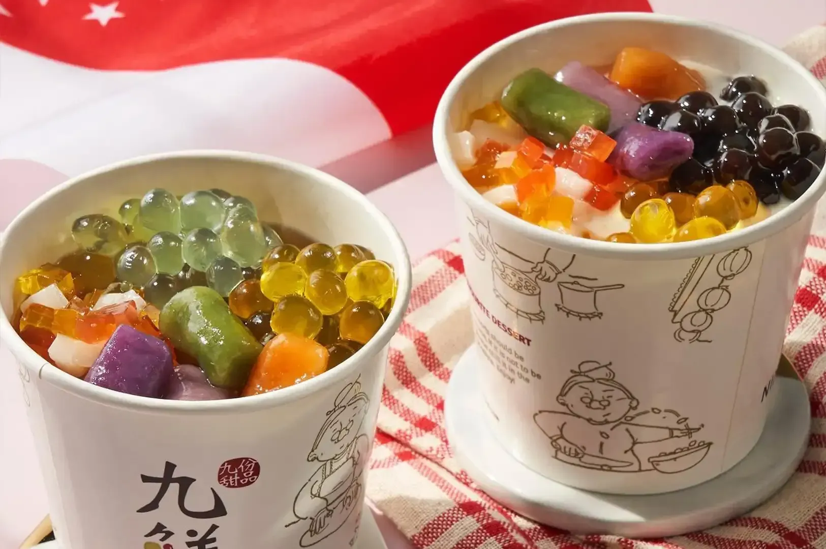 Two cups of shaved ice topped with colorful jelly and fruit on a red-and-white cloth