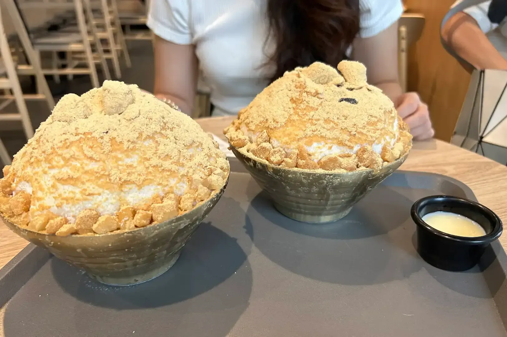 Two large bowls of shaved ice dessert on a table with a small cup of cream.