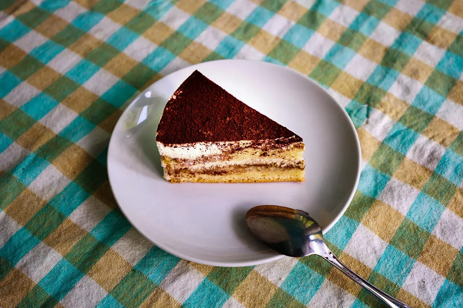 Slice of chocolate cake on a white plate with a spoon on a green and yellow checkered tablecloth