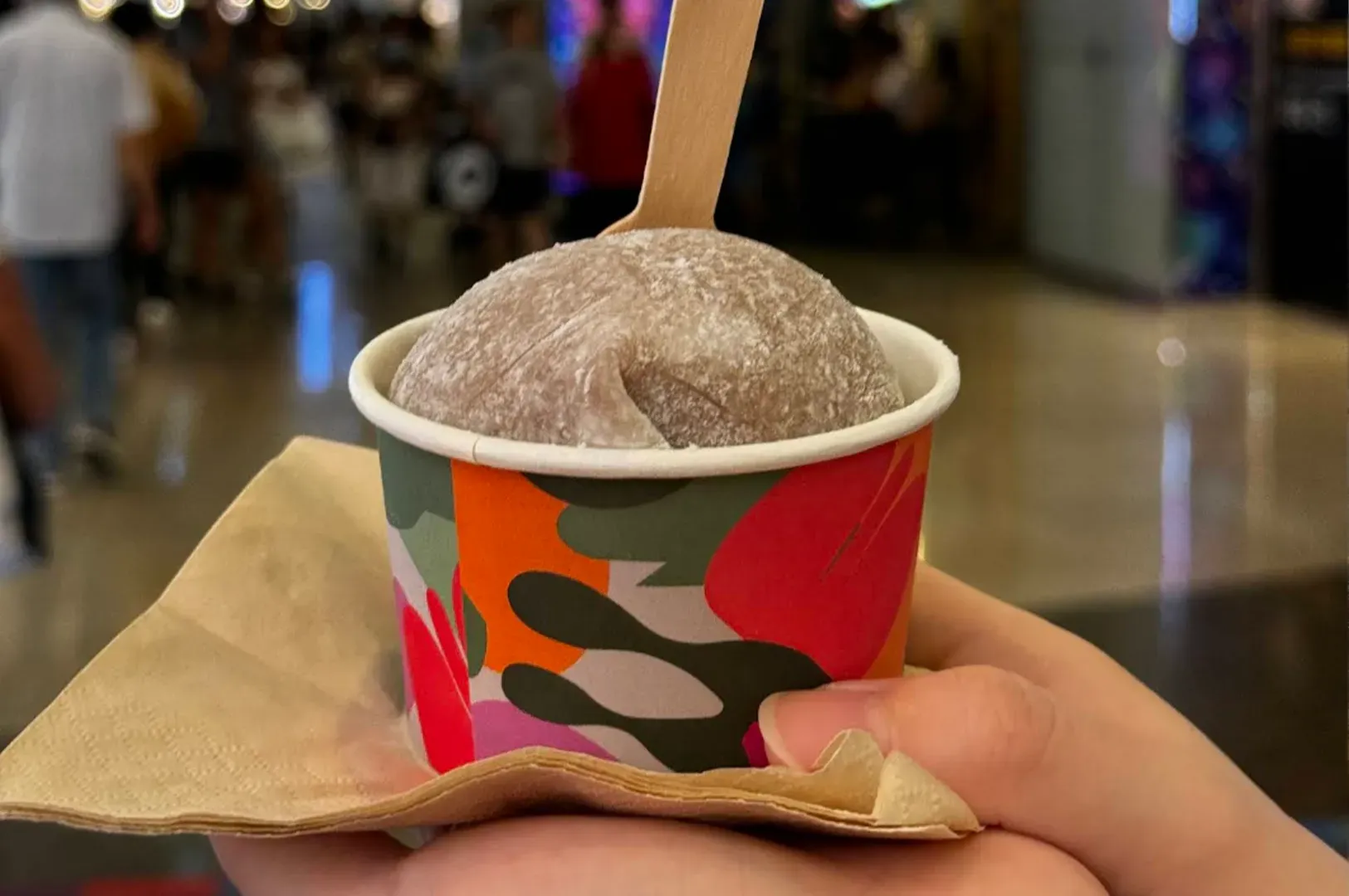 Cup of ice cream in a colorful paper bowl with a wooden spoon, held in a hand inside a mall.