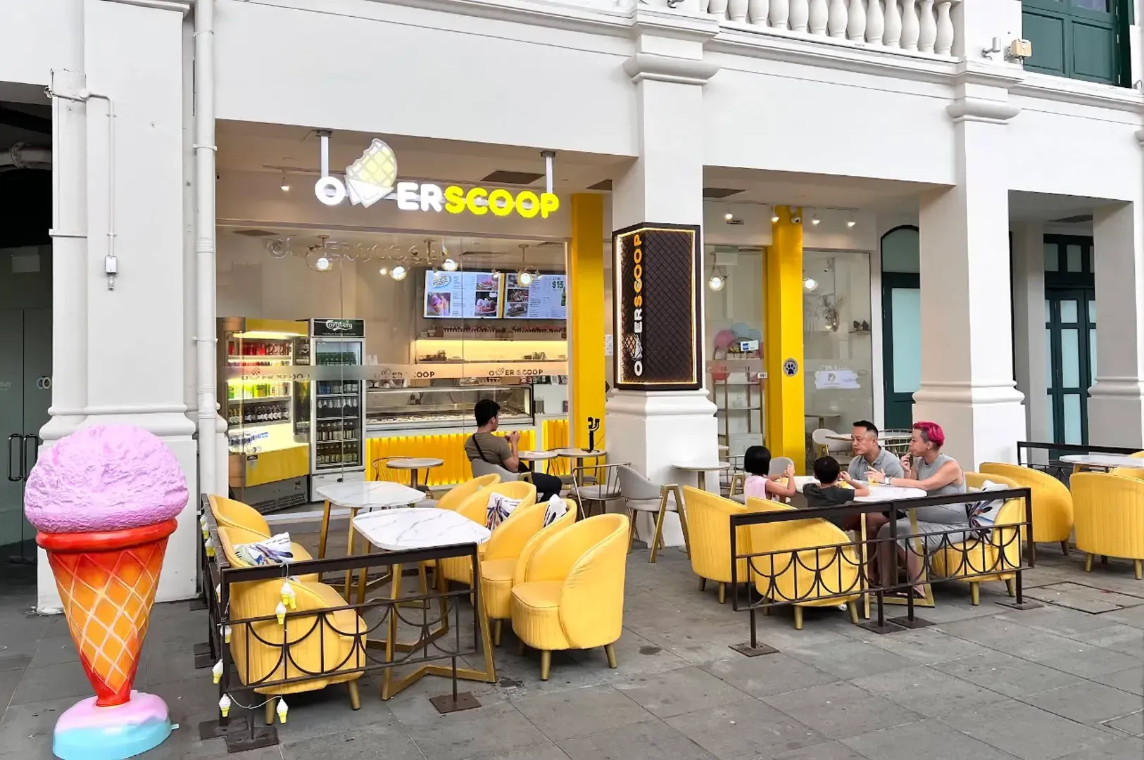 Outdoor café seating beside a yellow ice cream shop, with customers at tables and a pink cone sculpture.