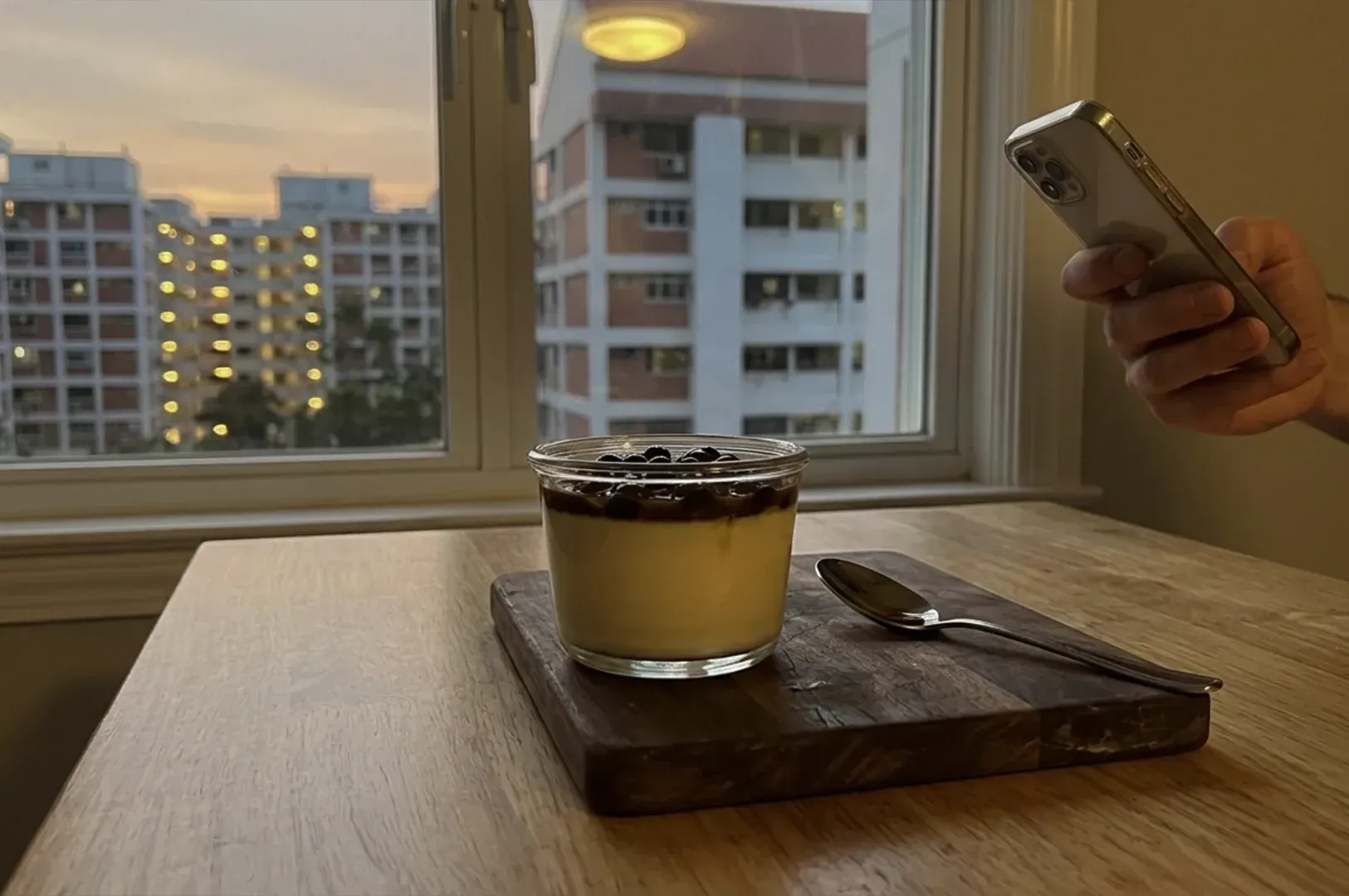 Dessert on a tray by a window, with a hand holding a phone and apartment buildings outside.