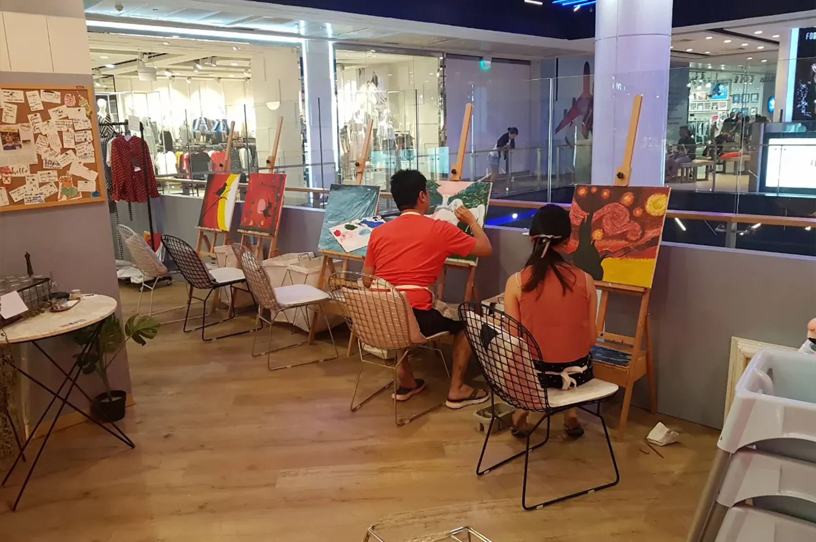 People seated on stools painting at easels in an indoor studio by a glass storefront.