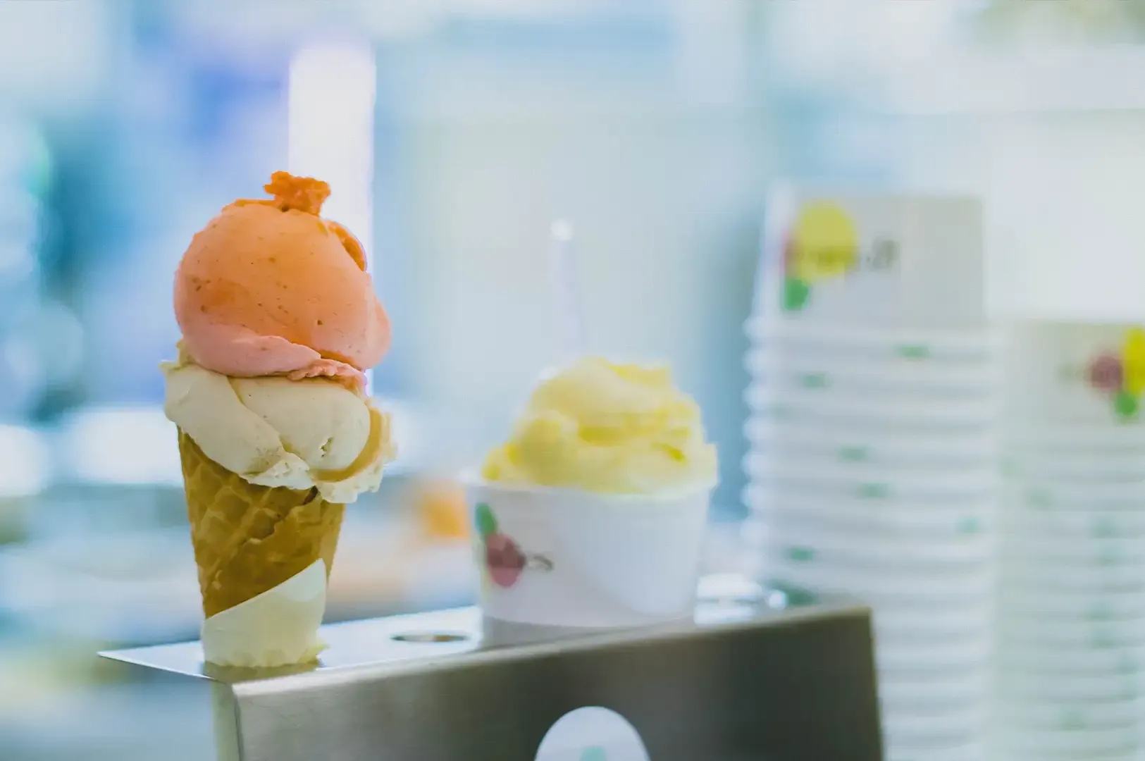 Two scoops of ice cream in a cone and cup on a counter, with stacked cups in the background